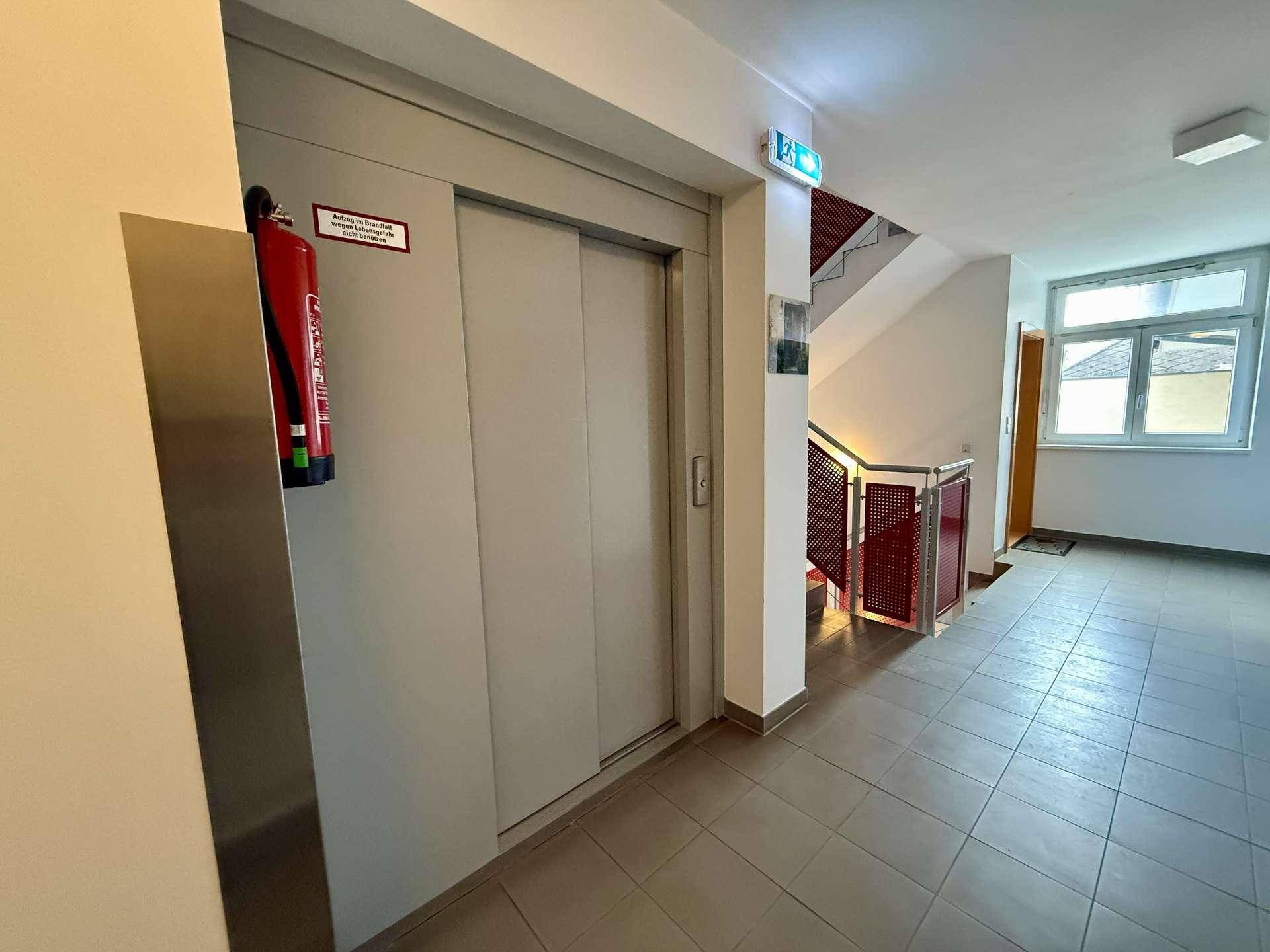 Heller Hausflur mit Aufzug, Treppenhaus und Fliesenboden im Eingangsbereich.