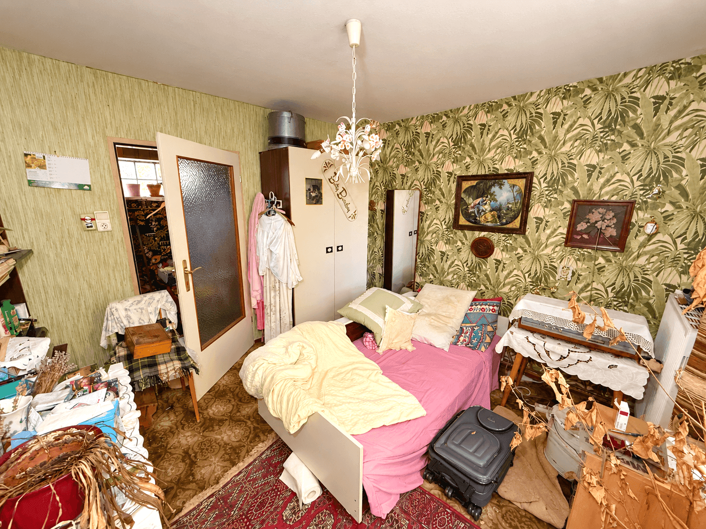 Schlafzimmer mit Einzelbett, grüner floraler Tapete und Holzdecke, viele Gegenstände.
