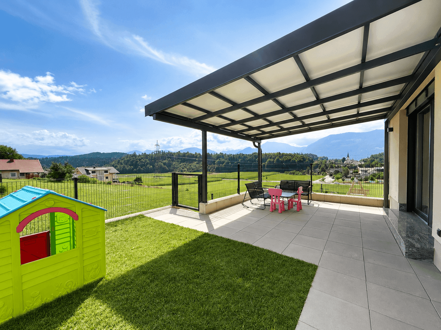 Überdachte Terrasse mit Sitzgelegenheiten, Spielhaus und weitem Blick über die grüne Landschaft.