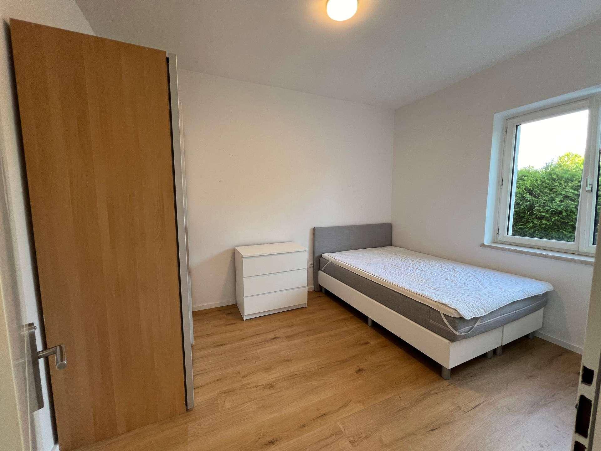 Helles Schlafzimmer mit Einzelbett, Kommode und großem Kleiderschrank, mit Blick ins Grüne.