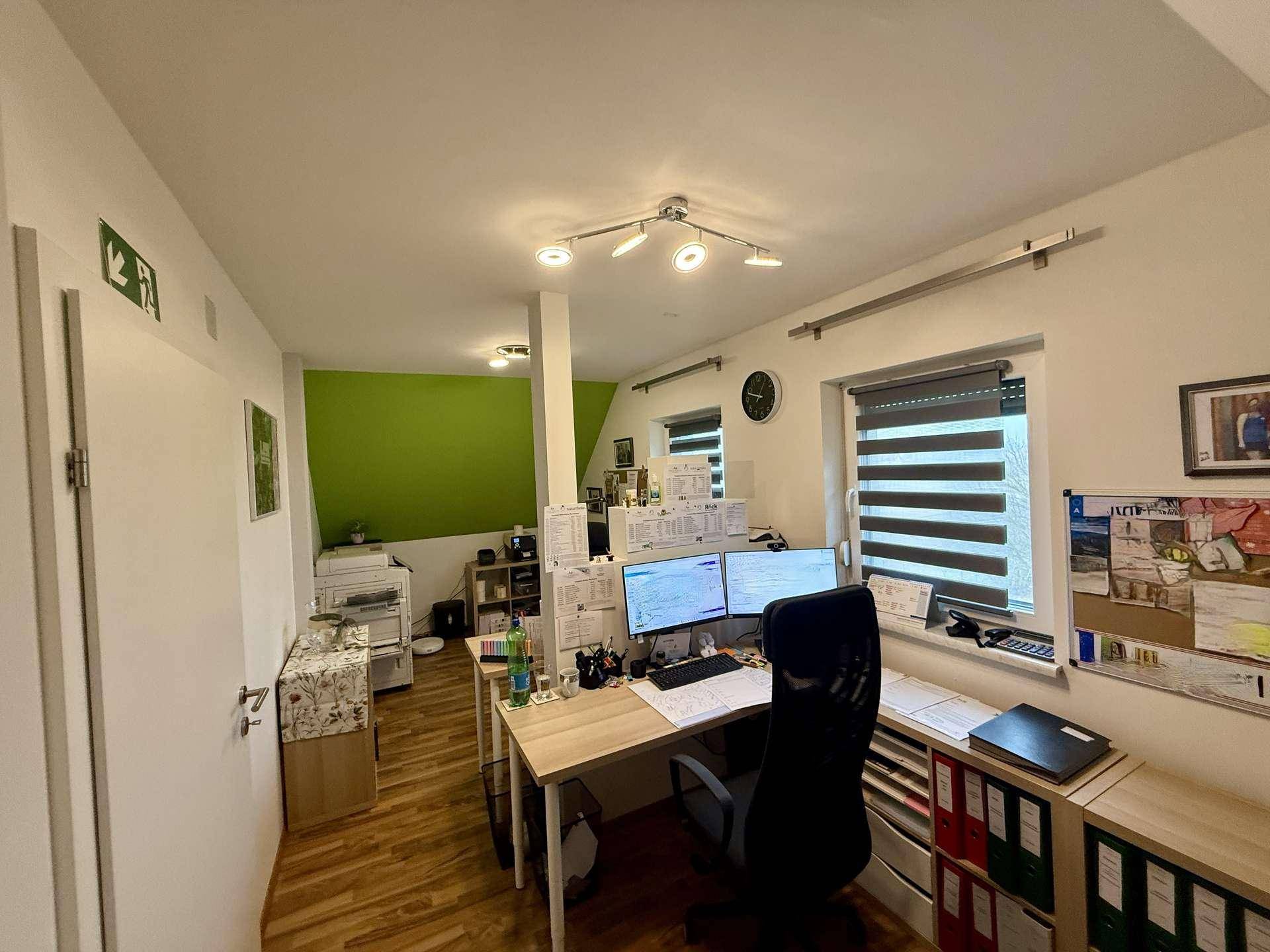 Helles Arbeitszimmer mit zwei Bildschirmen, Holzboden und einem Fenster mit Jalousien.