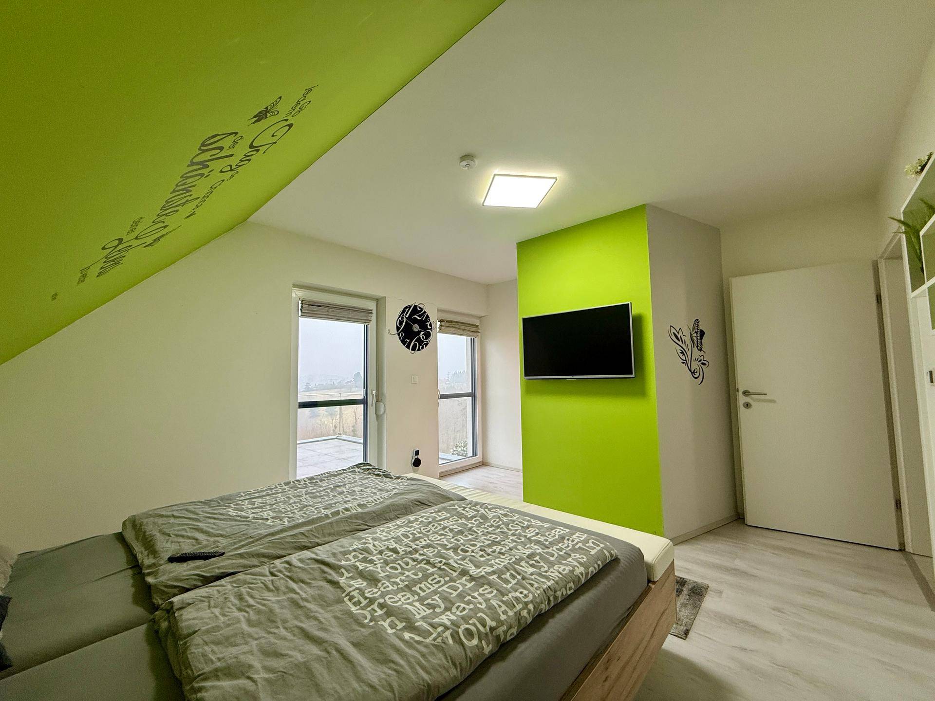 Modernes Schlafzimmer mit Doppelbett, grüner Akzentwand und Zugang zum Balkon.