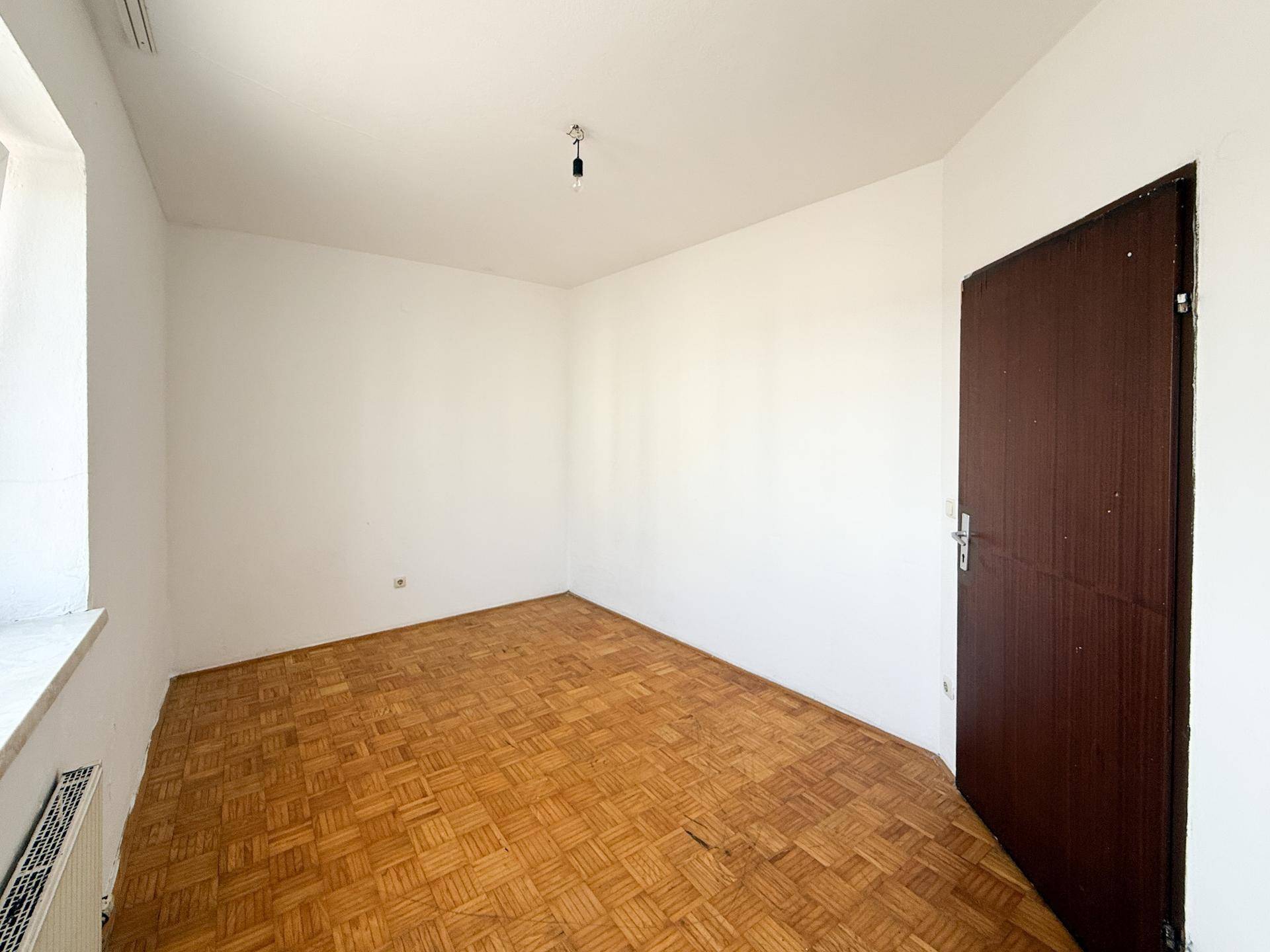 Leeres Zimmer mit Parkettboden und einer dunklen Holztür, ideal als Schlaf- oder Arbeitszimmer.