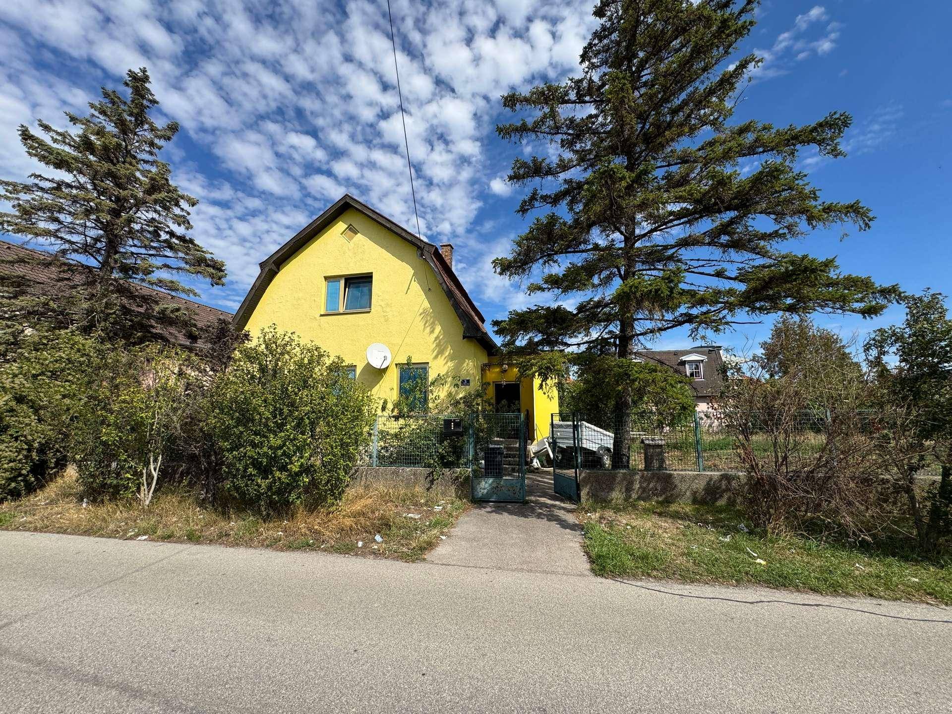 Gelbes Einfamilienhaus mit Satteldach, umgeben von Bäumen unter einem strahlend blauen Himmel.