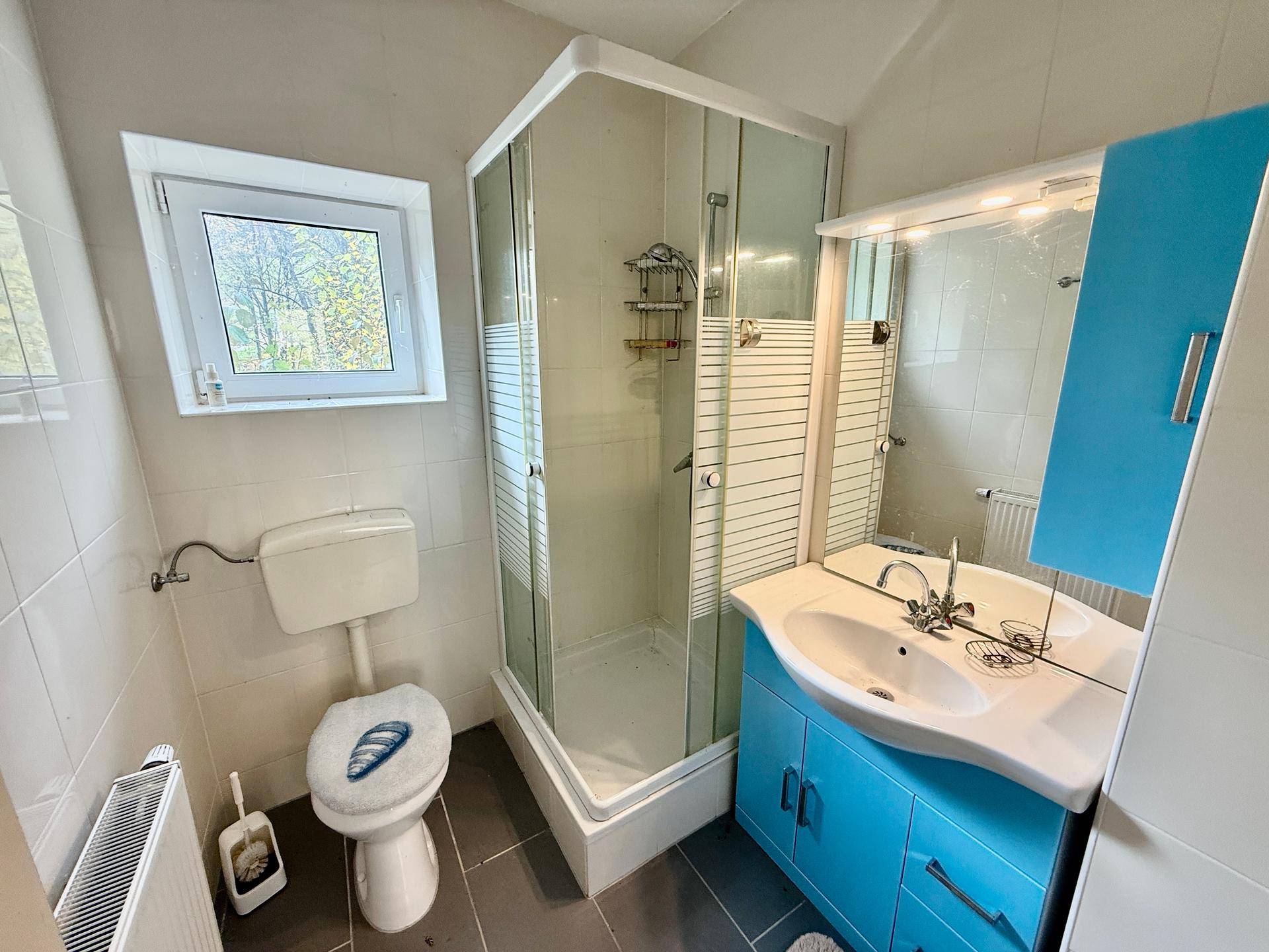 Modernes Badezimmer mit Duschkabine, WC und blauem Waschtischunterschrank, hell gefliest.