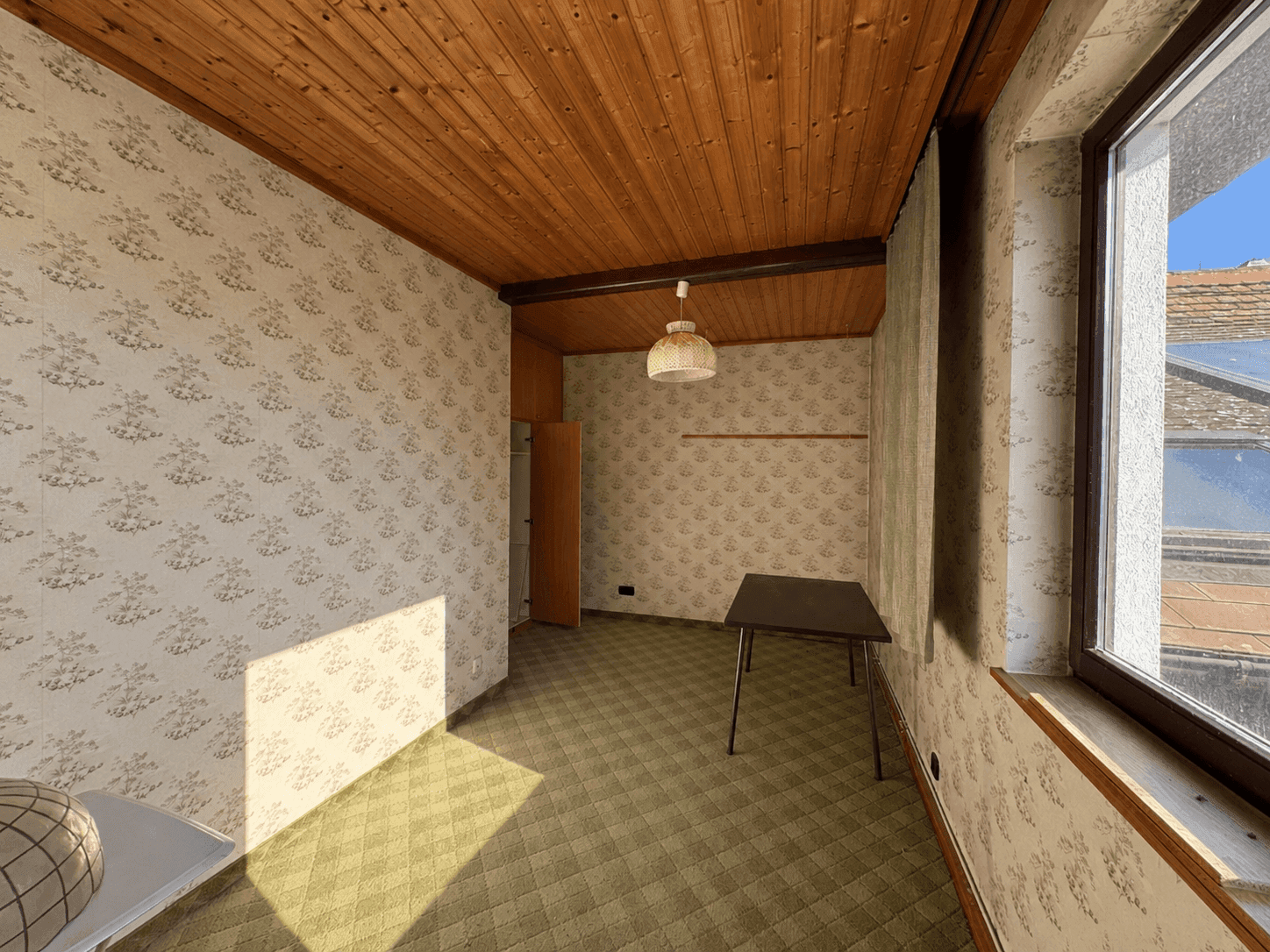 Kleines Zimmer mit Holzdecke, Teppichboden und Tapeten, hell durch großes Fenster.