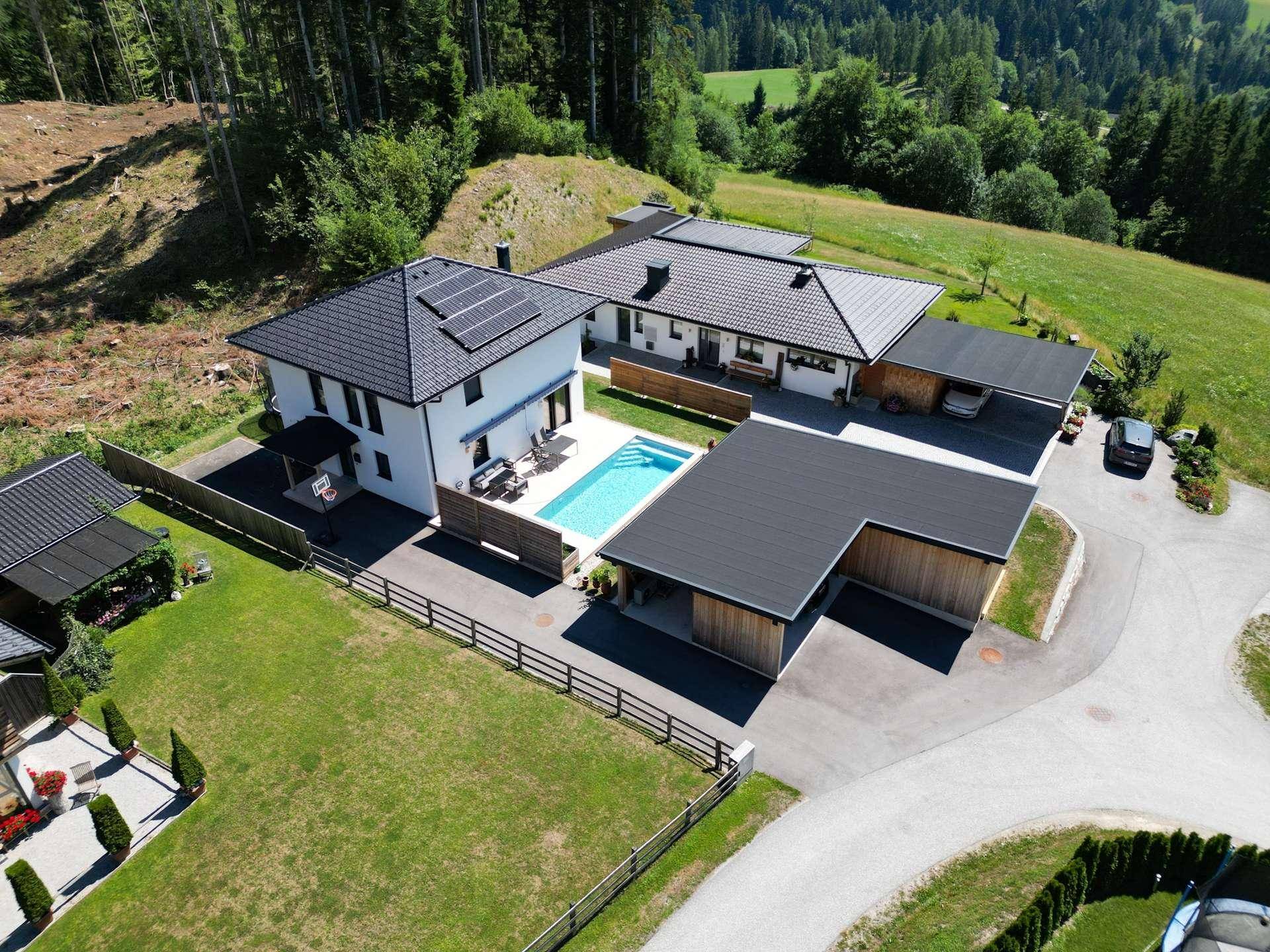 Weitläufige Luftaufnahme des Anwesens mit zwei Häusern, Pool und Carport, umgeben von Wald und Bergen.
