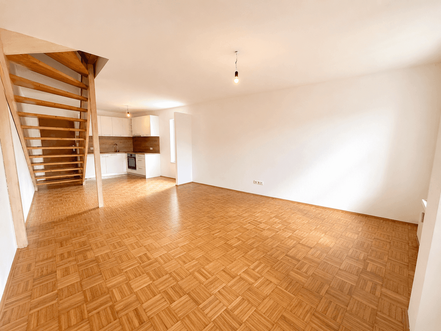Weitläufiger Wohnbereich mit Parkettboden, offener Küche und Holztreppe zum oberen Stockwerk.