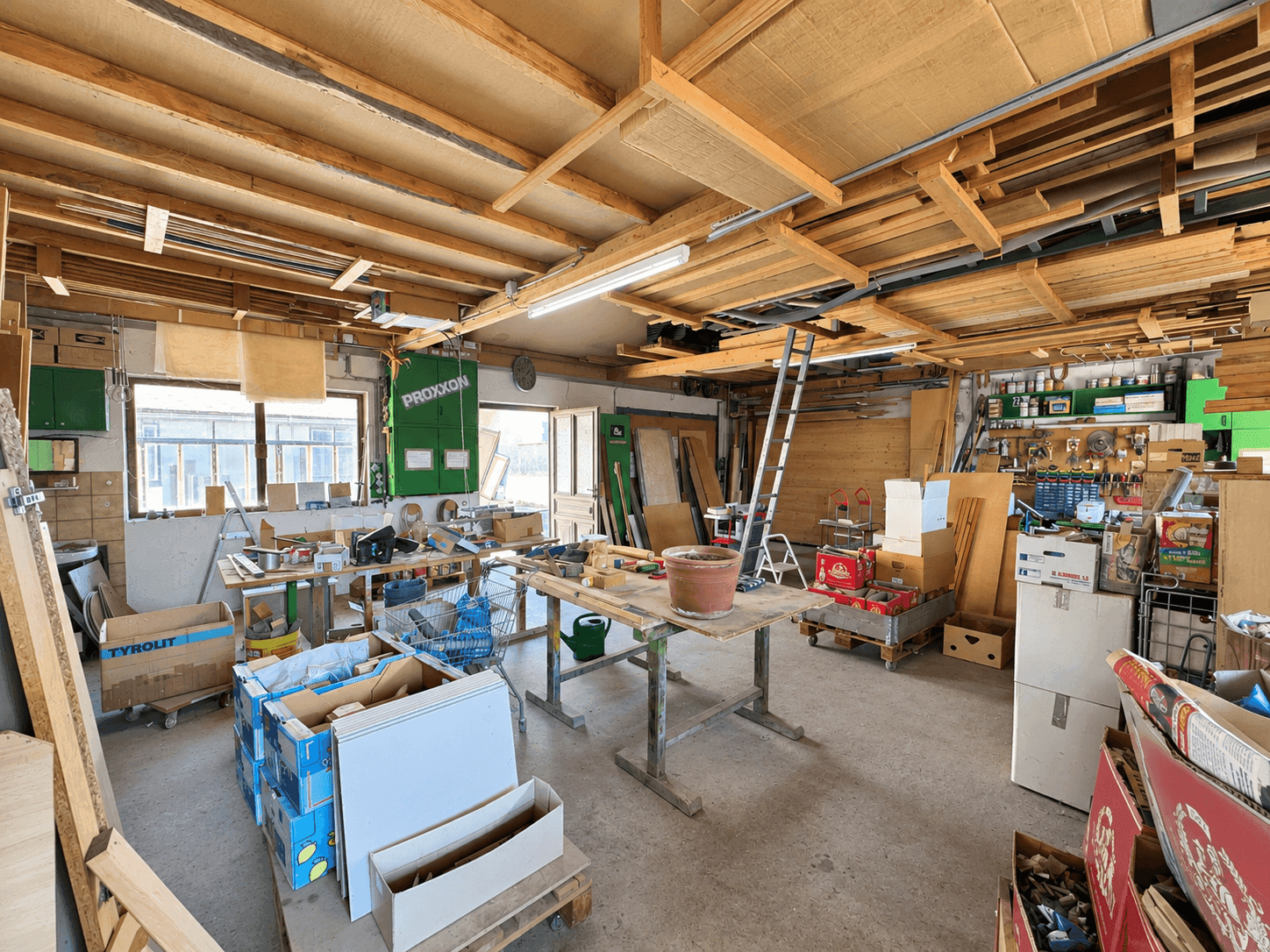 Große Werkstatt mit vielen Holzmaterialien, Werkbänken und Fenstern.