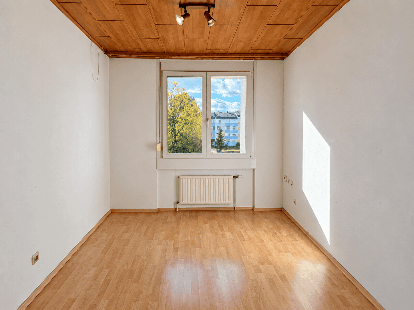 Helles Zimmer mit Laminatboden, Holzdecke und Fenster mit Blick auf die umliegende Wohnbebauung.