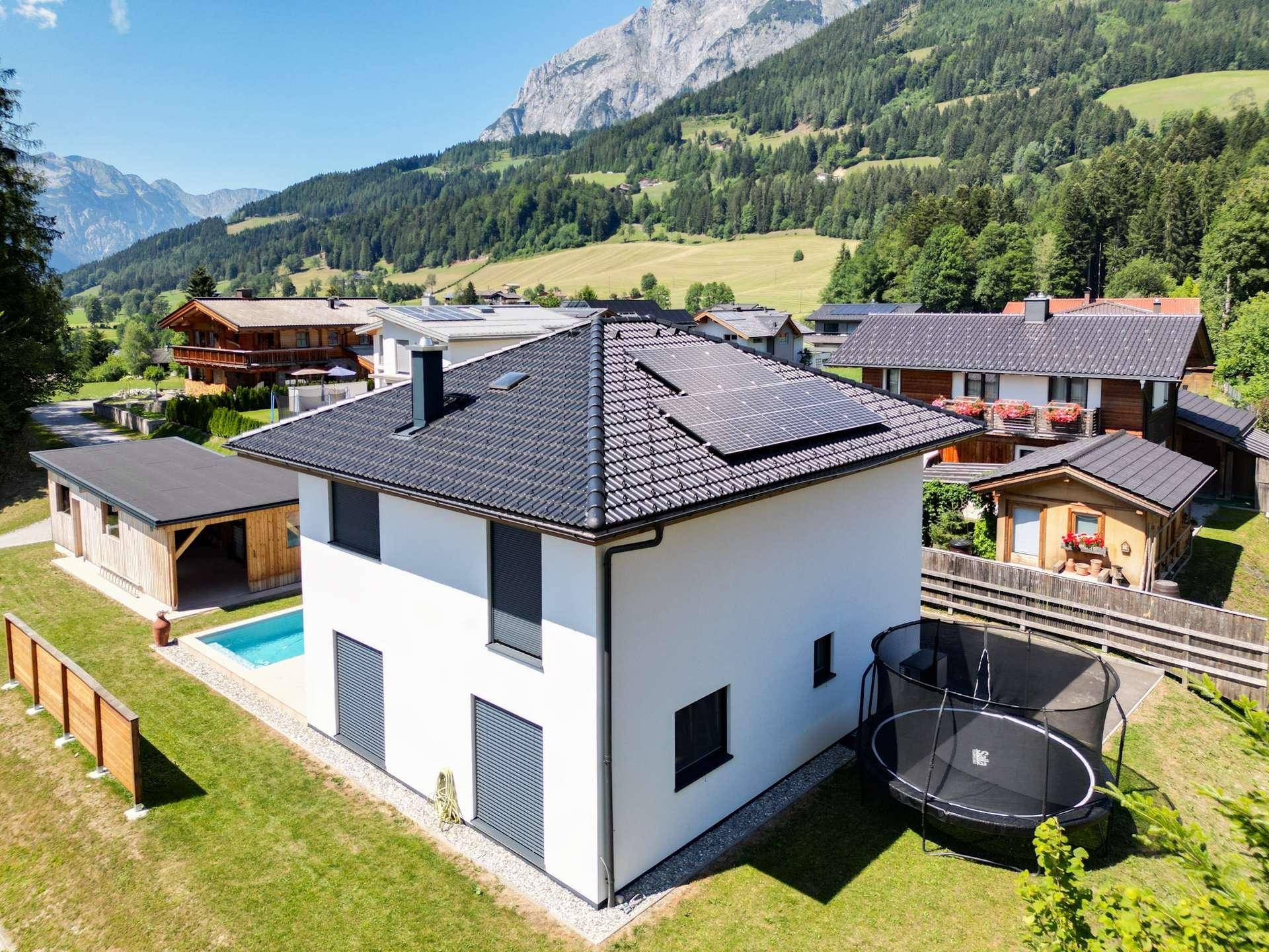 Modernes weißes Haus mit Pool und Trampolin im Garten, vor einer beeindruckenden Bergkulisse.