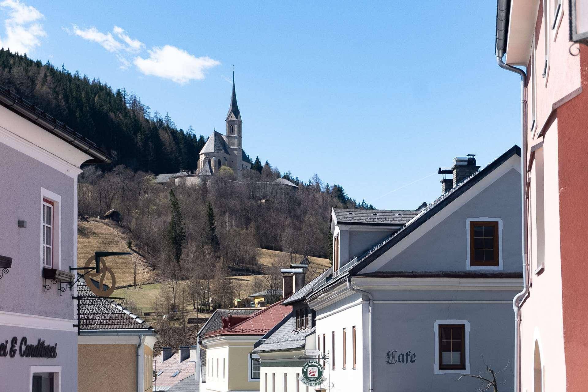 Malerische Dorfansicht mit einer Kirche auf einem bewaldeten Hügel unter blauem Himmel.