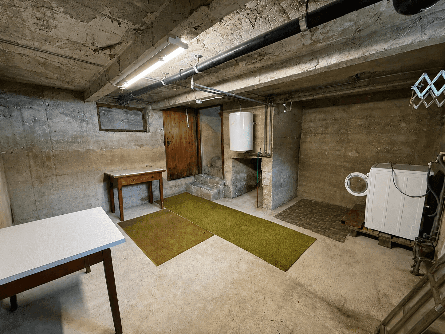 Kellerraum mit Waschmaschine, Warmwasserboiler, Betonwänden und sichtbaren Rohren.