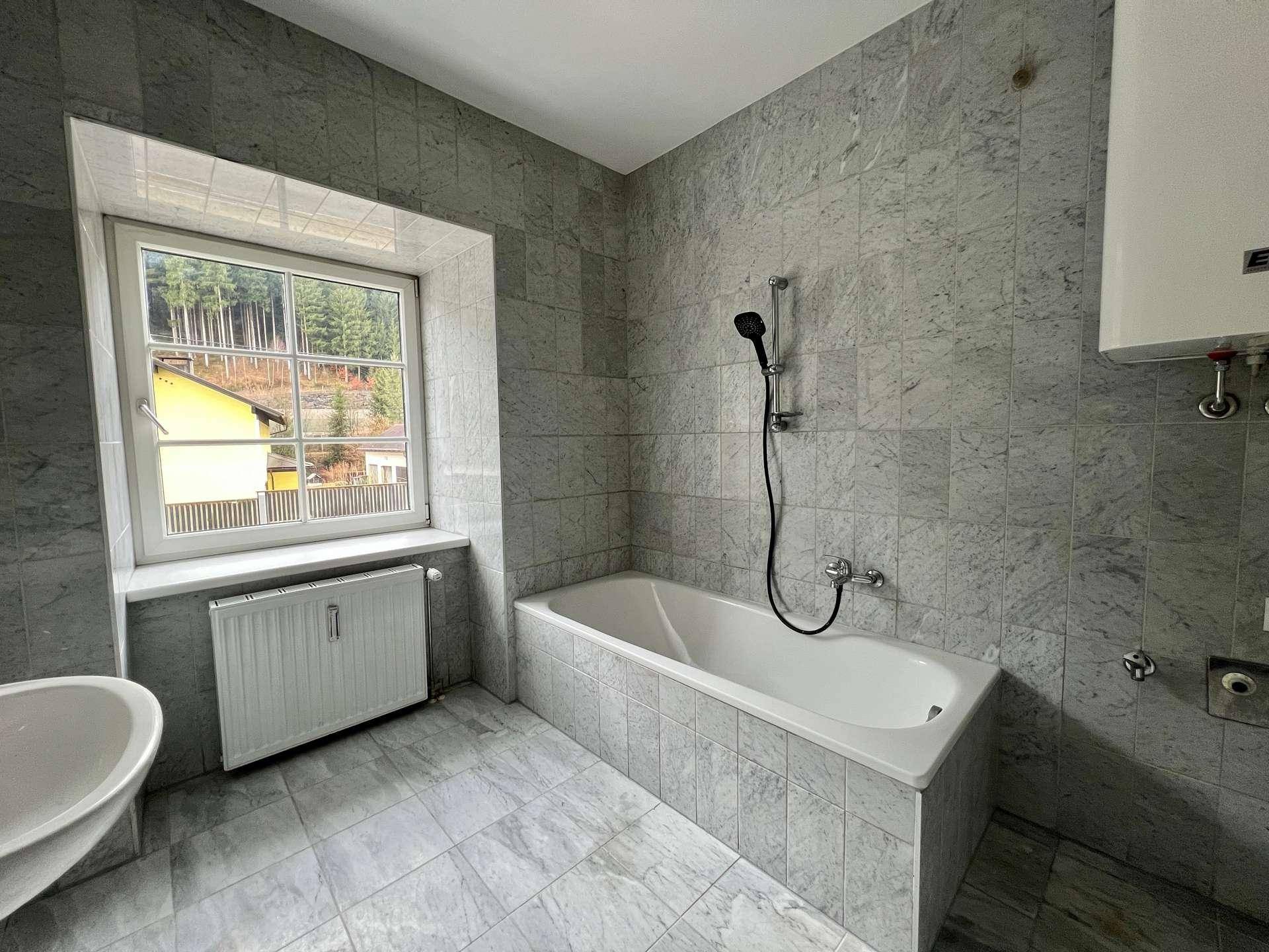 Badezimmer mit grauen Fliesen, Badewanne und Fenster mit Blick auf bewaldete Hügel.