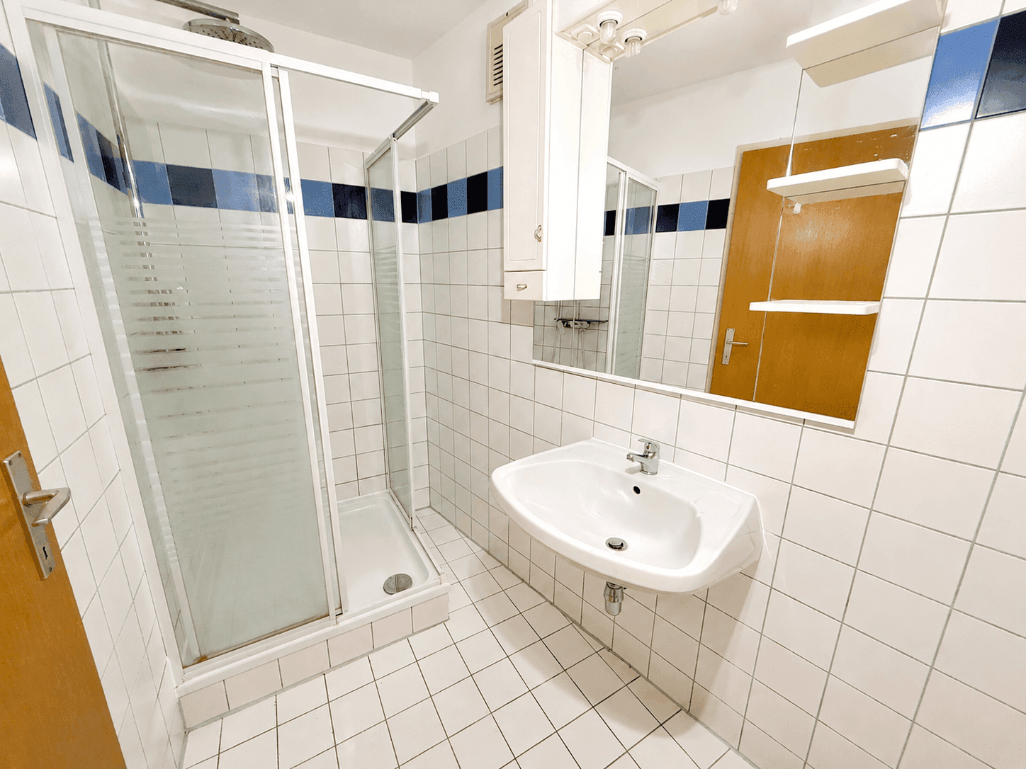 Badezimmer mit Duschkabine, Waschbecken, Spiegel und weißen Fliesen mit blauen Akzenten.