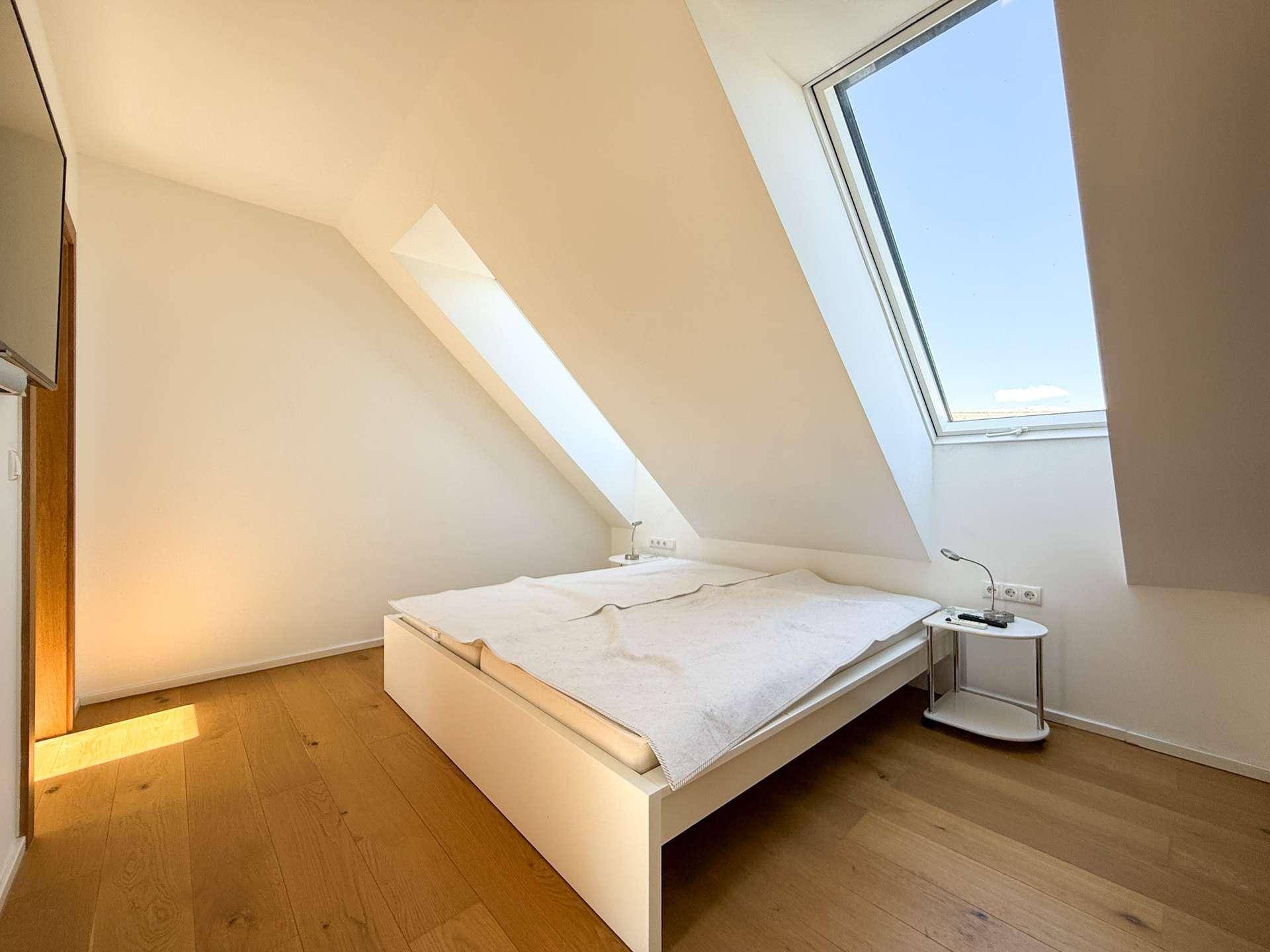 Helles Schlafzimmer mit Holzboden, weißem Bett und zwei Dachfenstern, die den Raum mit Licht füllen.