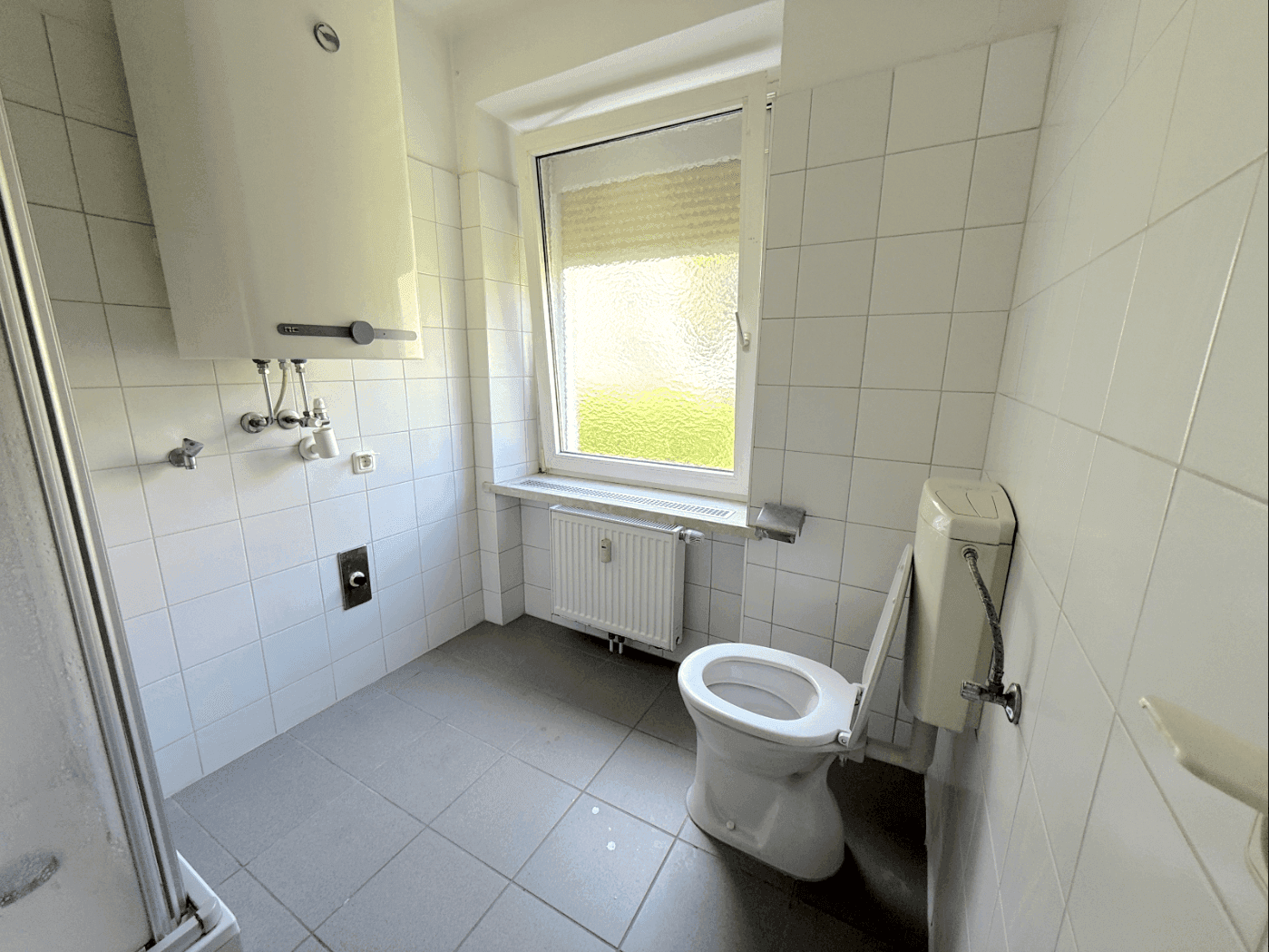 Einfaches Badezimmer mit weißen Fliesen, Toilette, Dusche und einem Fenster.