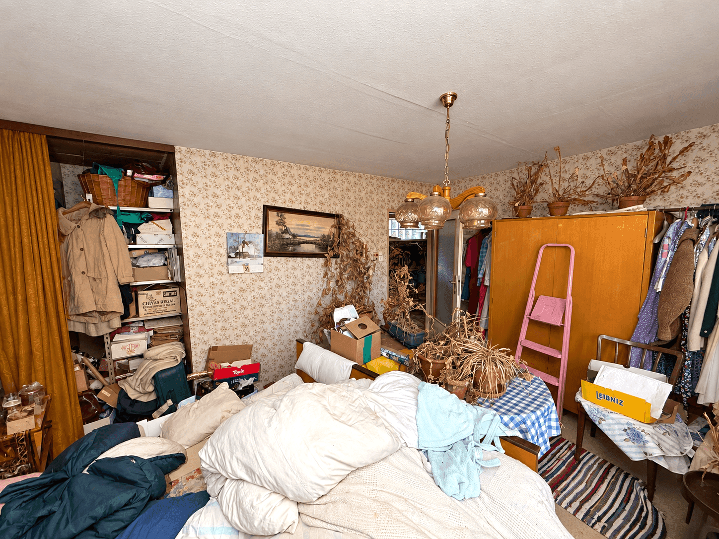 Schlafzimmer mit Bett, floraler Tapete und offenem Kleiderschrank, unaufgeräumt.