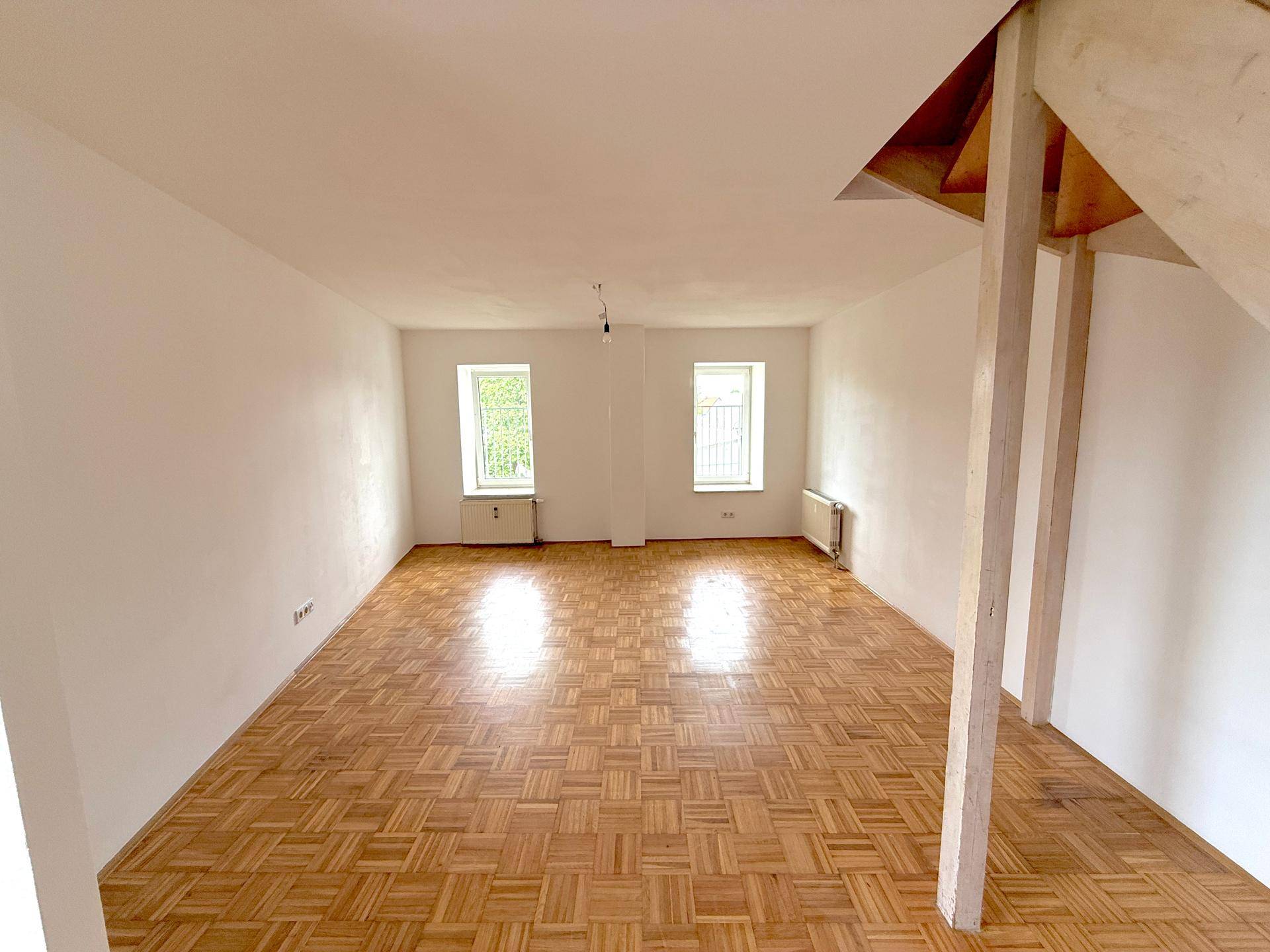 Geräumiger, heller Wohnraum mit Parkettboden und zwei Fenstern, offen gestaltet.