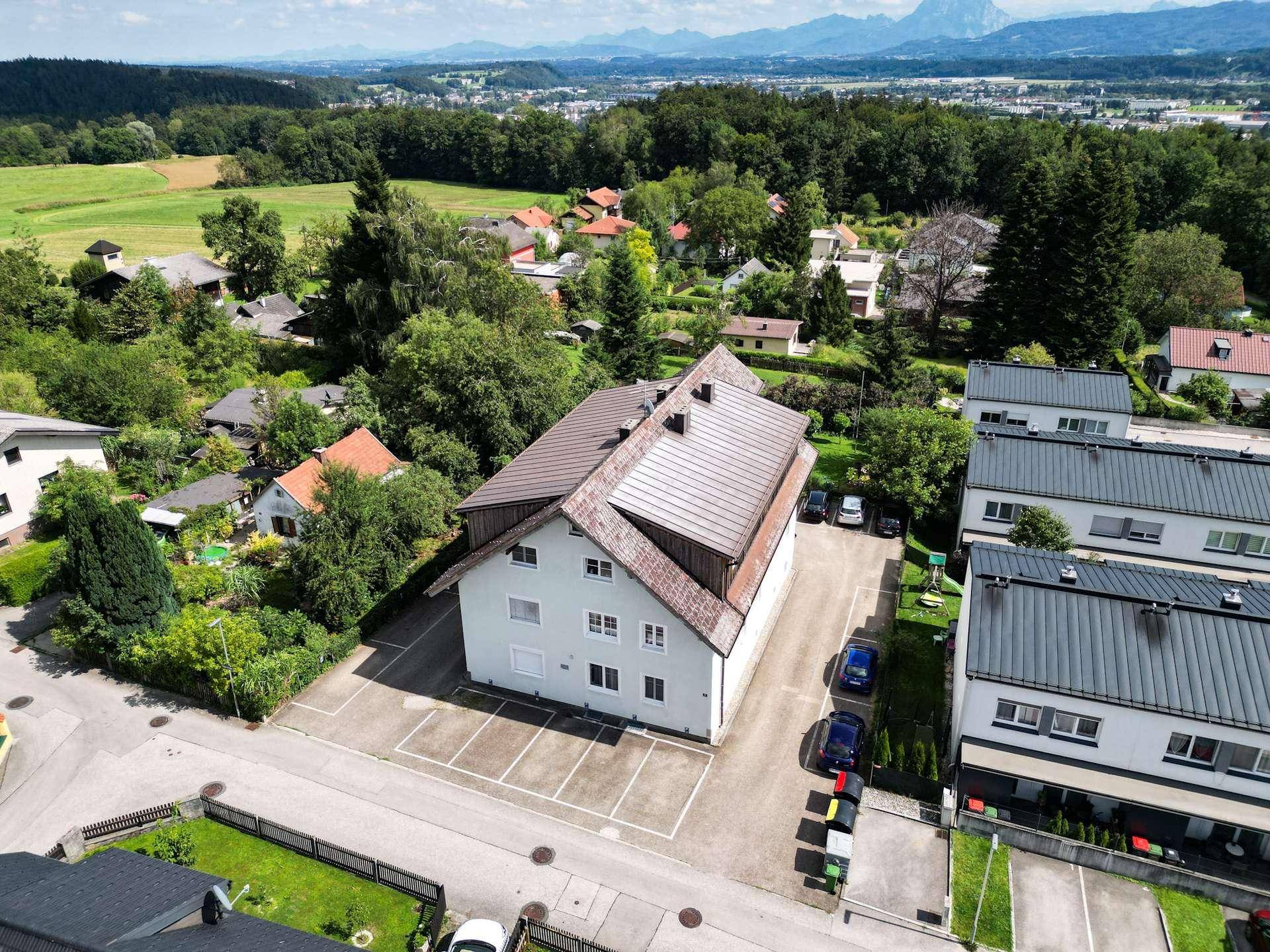Luftaufnahme eines Wohngebäudes mit Parkplätzen, umgeben von Grünflächen und Bergen im Hintergrund.