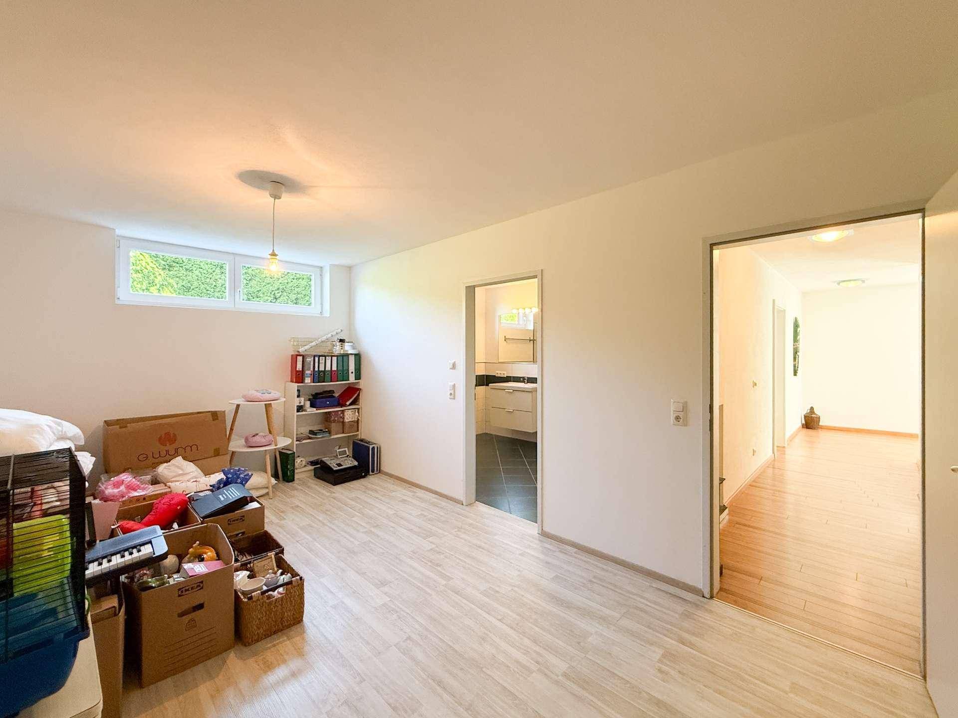 Ausgebauter Kellerraum mit hellem Boden, Fenster und Zugang zu einem weiteren Raum.