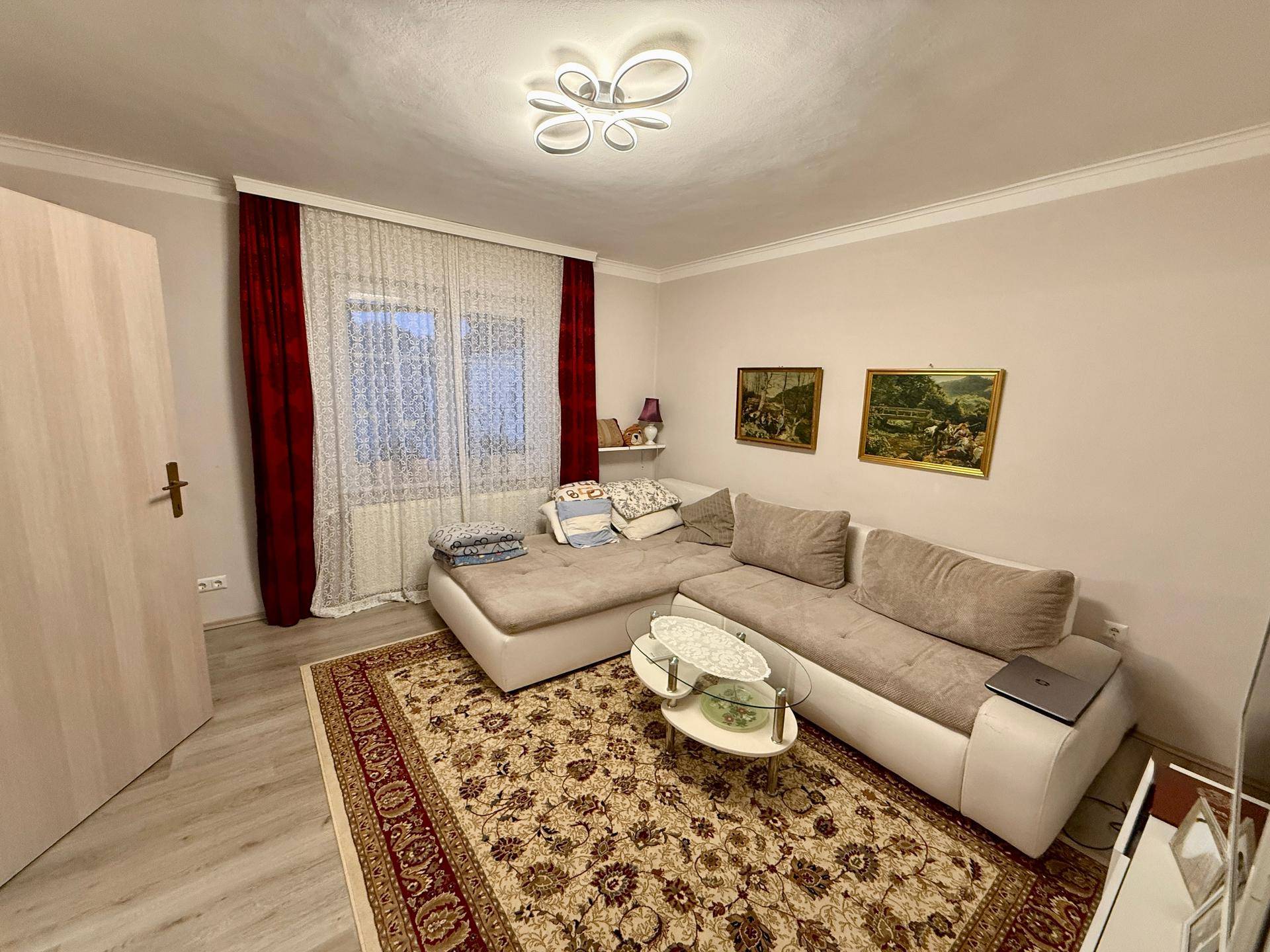 Helles Wohnzimmer mit beigem Ecksofa, gemustertem Teppich und Fenster mit roten Vorhängen.