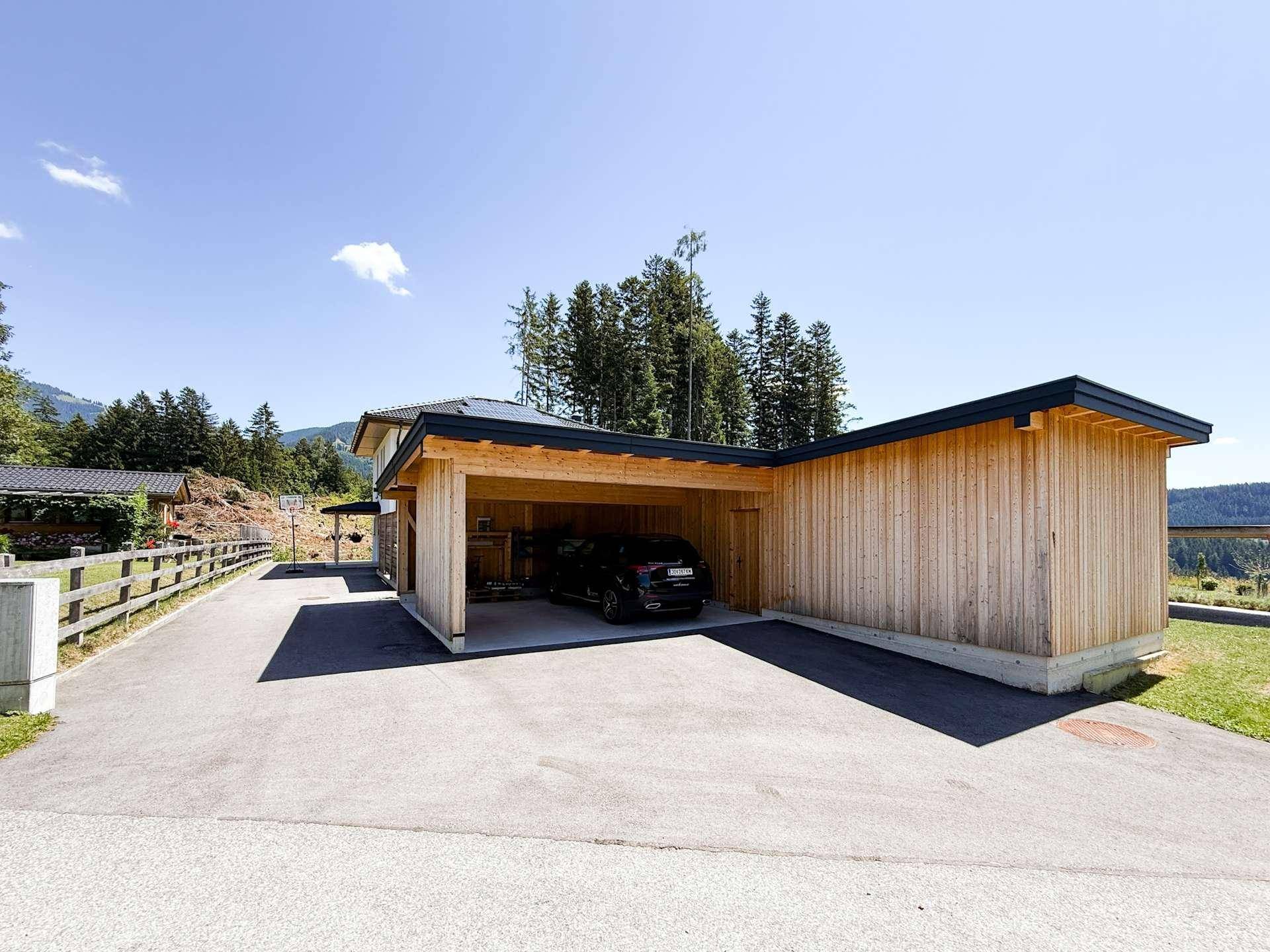 Offener Carport aus Holz mit Zufahrt und umgeben von grüner Landschaft.