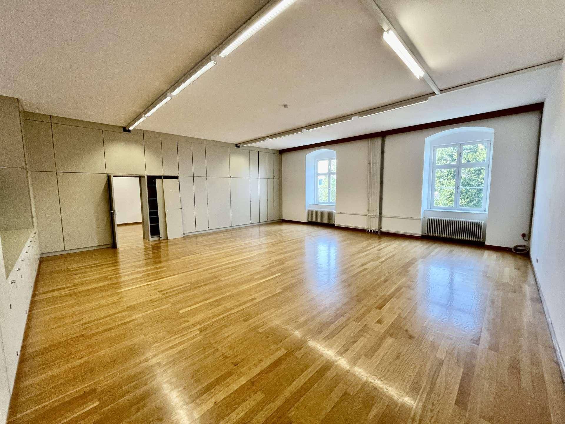 Geräumiger Raum mit Holzboden, zwei Fenstern und einer langen Wand mit integrierten Schränken.