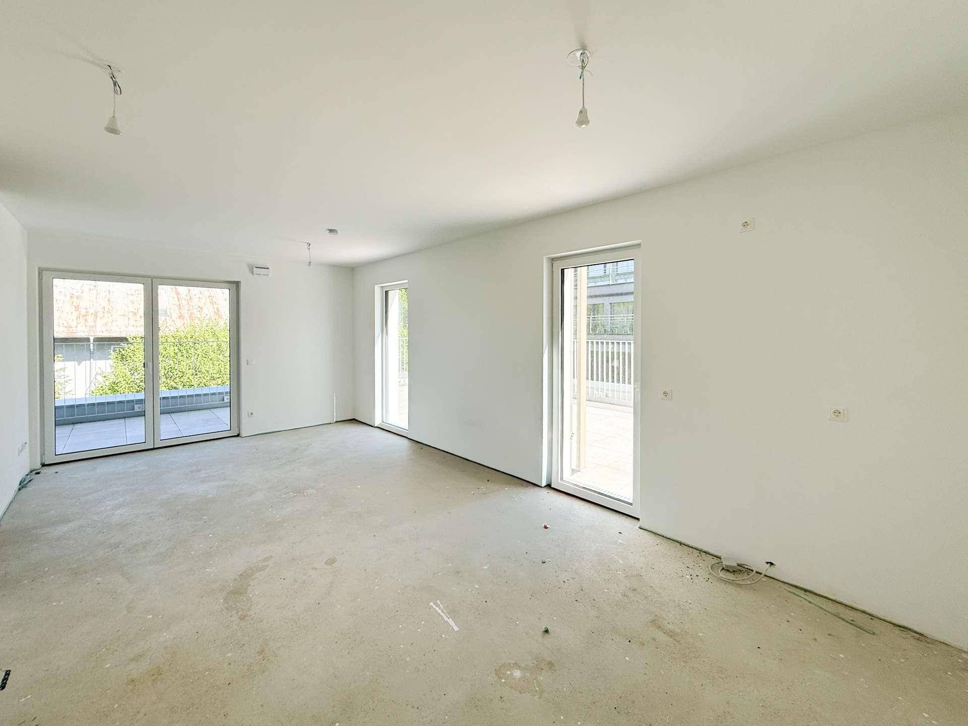 Unmöblierter Raum im Rohbauzustand mit Betonboden und großem Fenster zur Terrasse.