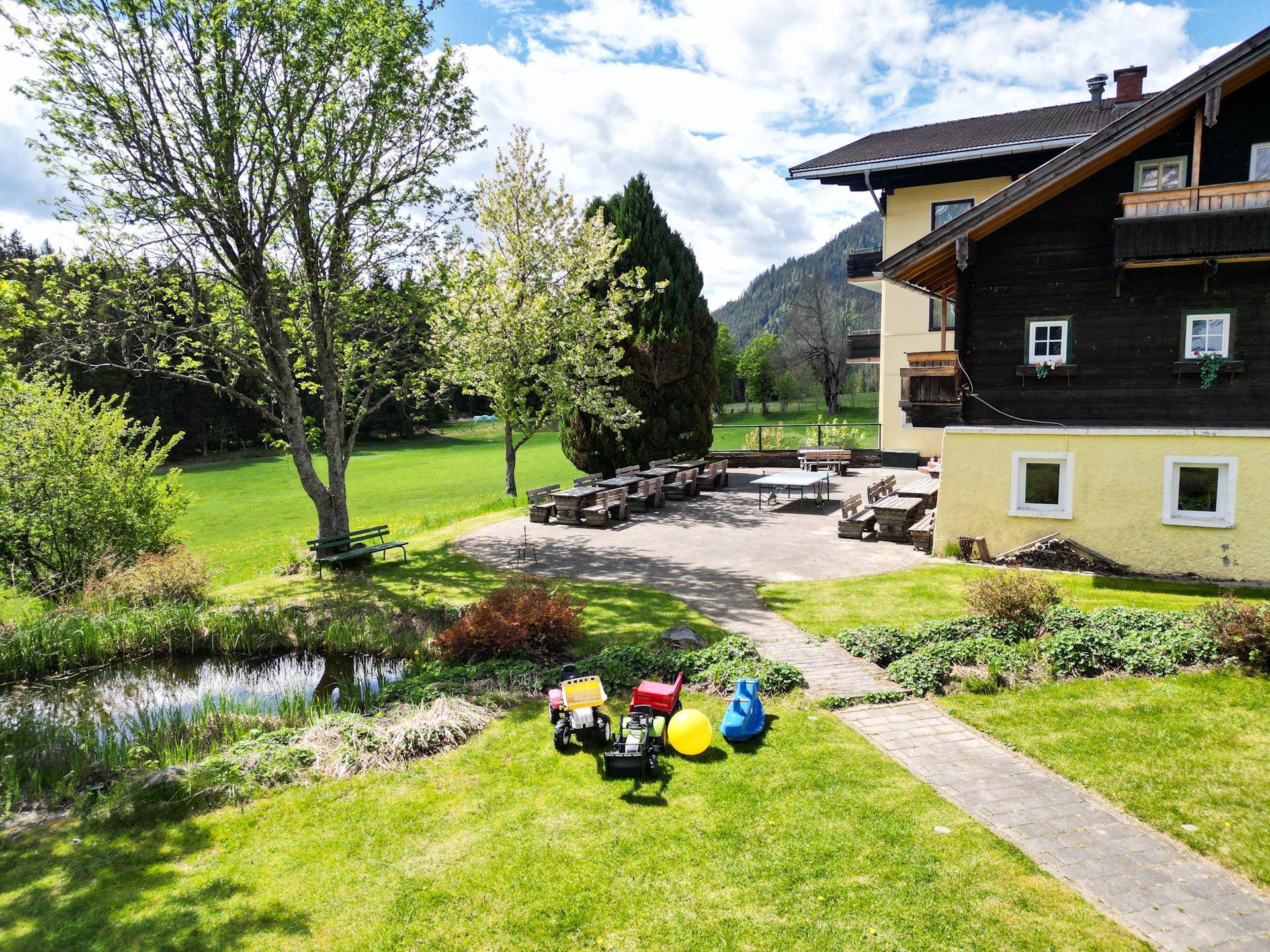 Gartenbereich mit Terrasse, Teich und Kinderspielzeug neben einem traditionellen Gebäude.