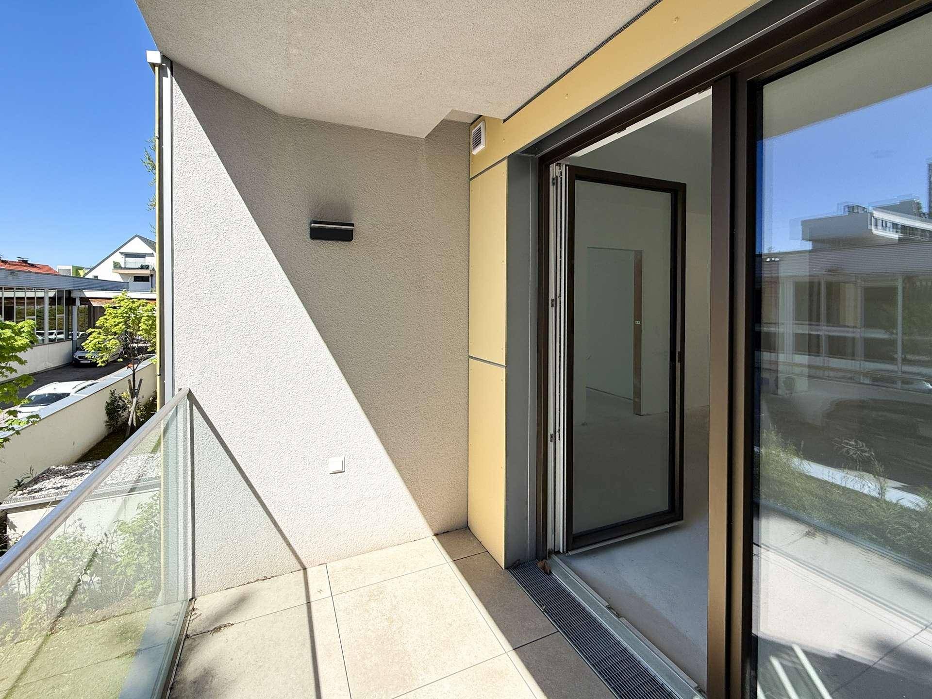 Sonniger Balkon mit Glasgeländer und offener Tür, die den Blick in den Innenraum freigibt.