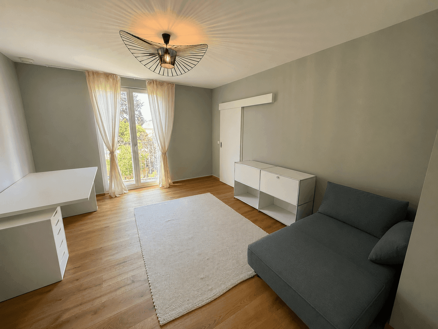 Helles Schlafzimmer mit Holzboden, Schreibtisch, Sofa und Zugang zu einem französischen Balkon.