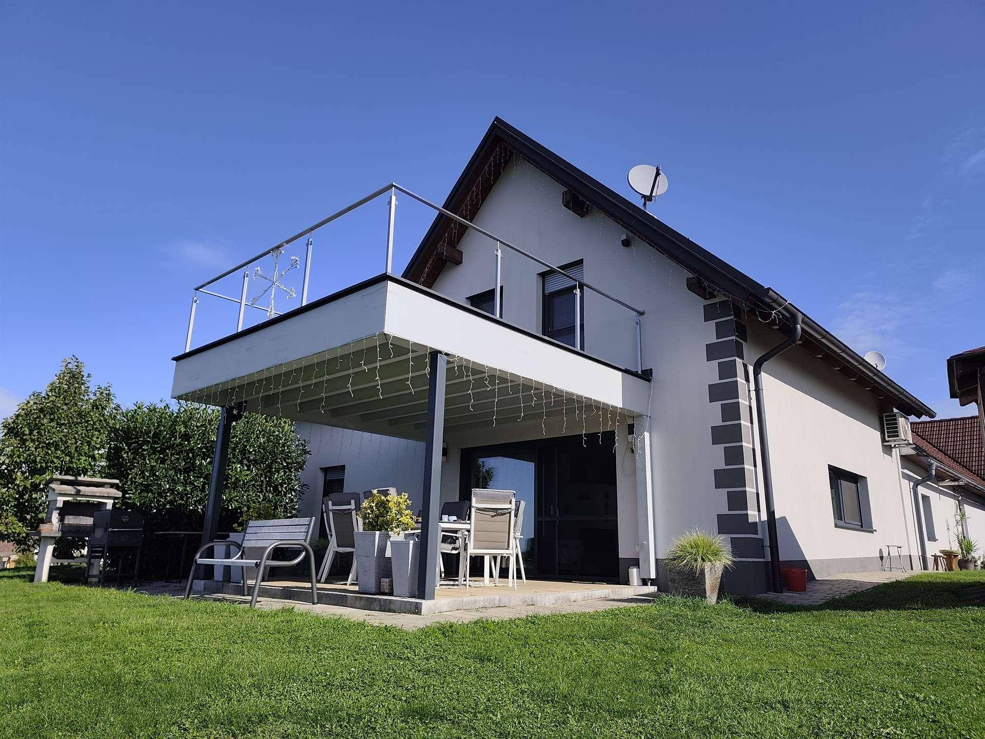 Terrasse mit Sitzgelegenheiten und Balkon darüber, umgeben von Grünflächen.