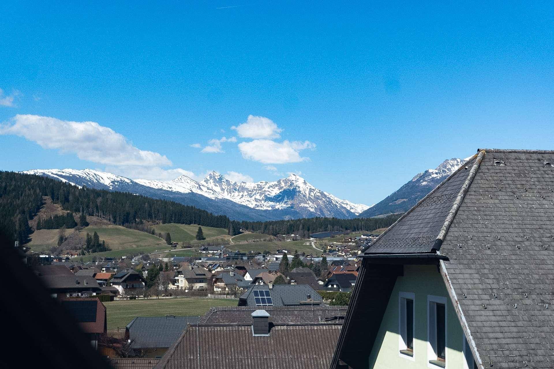 Panoramablick über eine malerische Siedlung mit schneebedeckten Bergen im Hintergrund unter blauem Himmel.