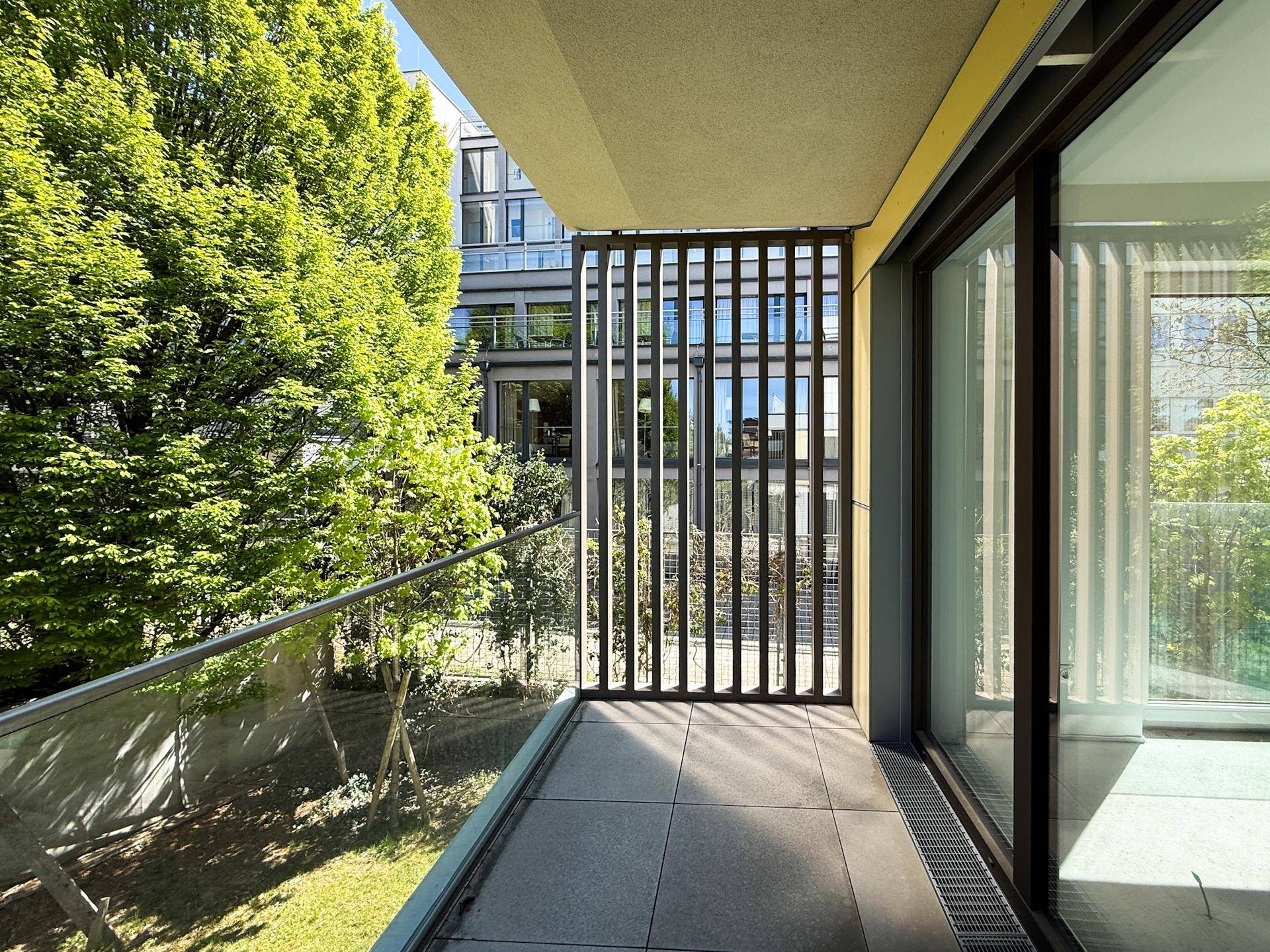 Moderner Balkon mit Glasgeländer, Fliesenboden und Blick auf Bäume und Gebäude.