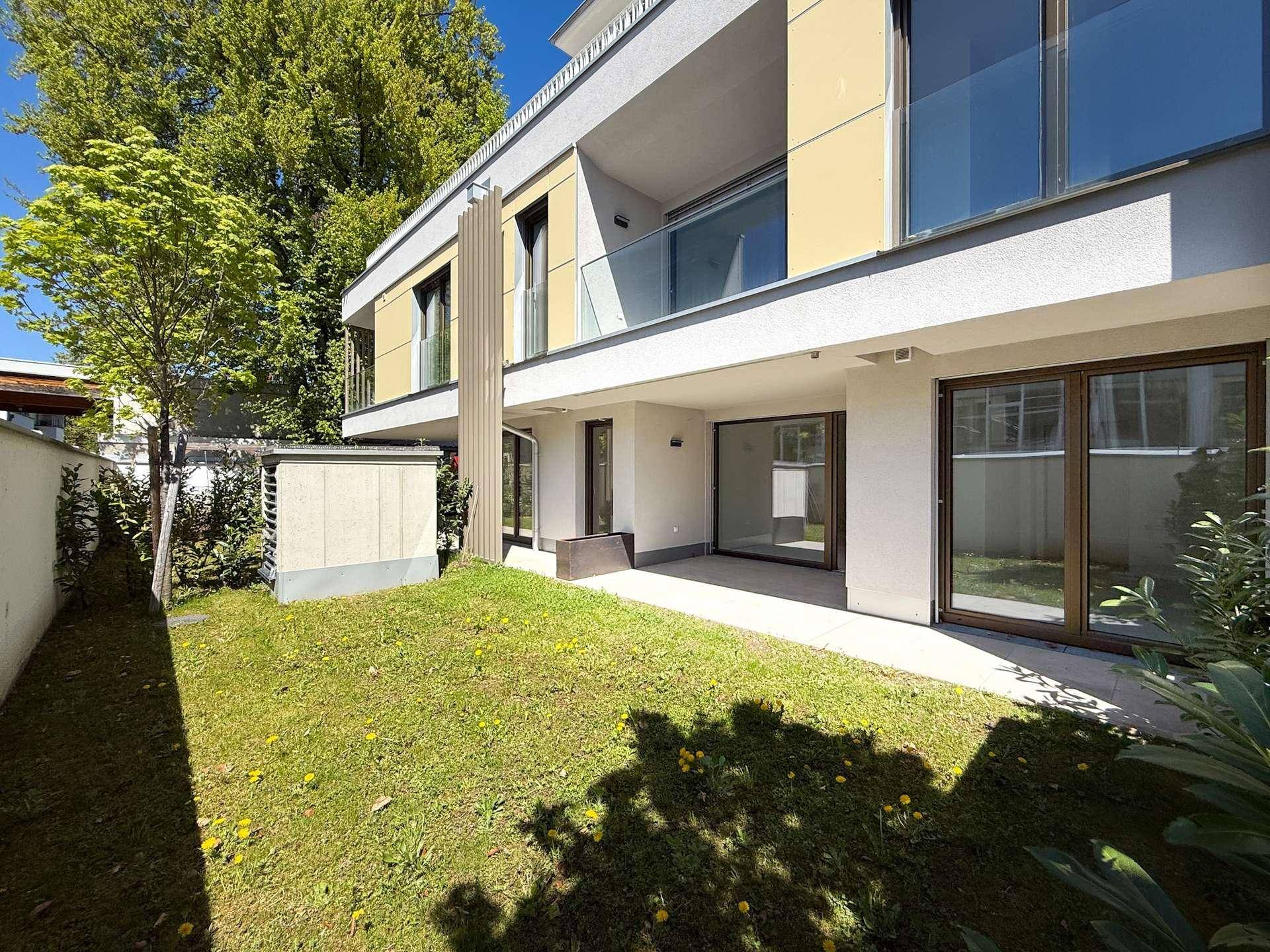 Gepflegter Gartenbereich mit Terrasse vor einer modernen Wohnanlage bei Sonnenschein.
