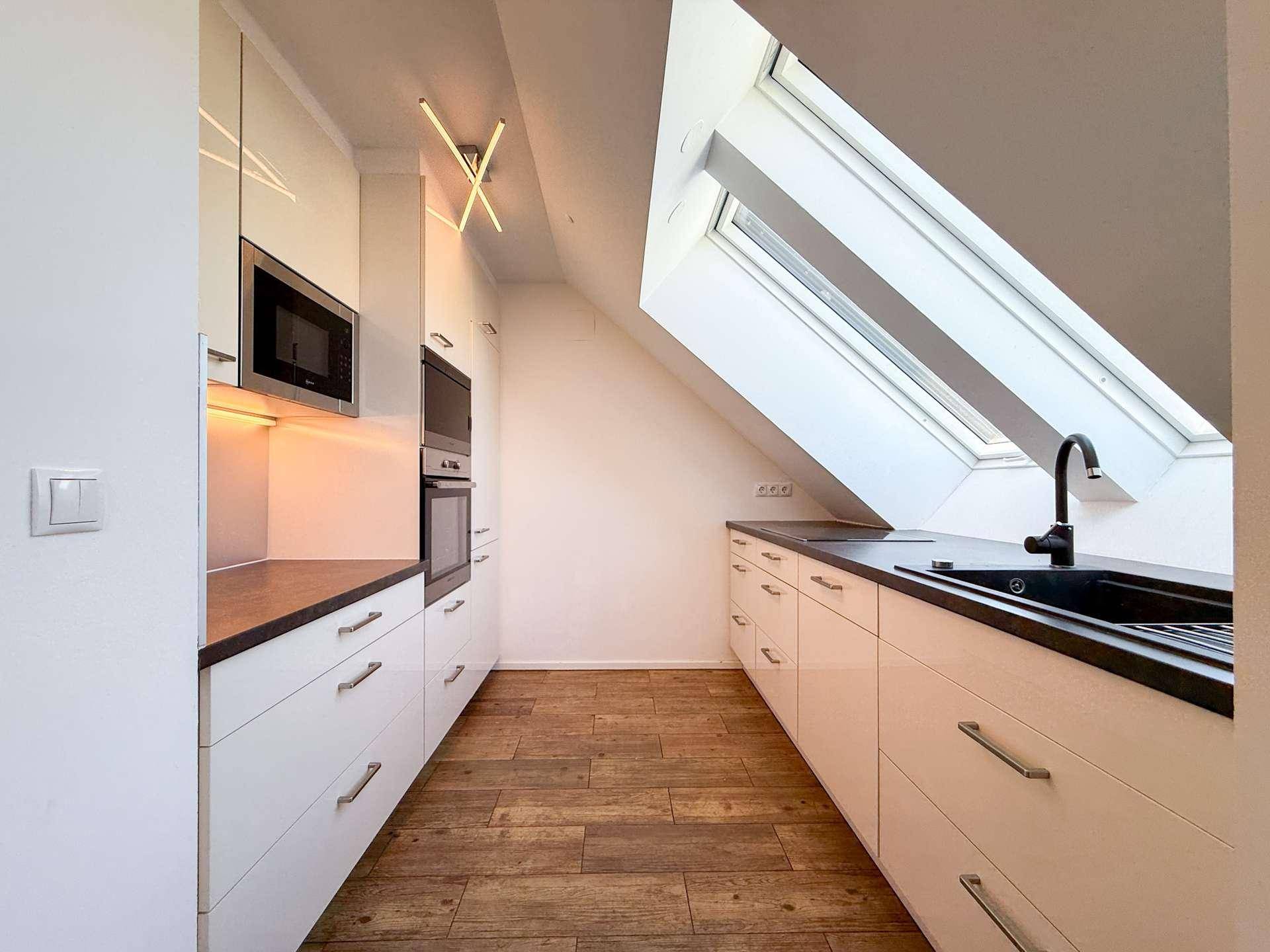 Moderne Küche mit weißen Schränken, dunklen Arbeitsplatten, Einbaugeräten und einem hellen Dachfenster.