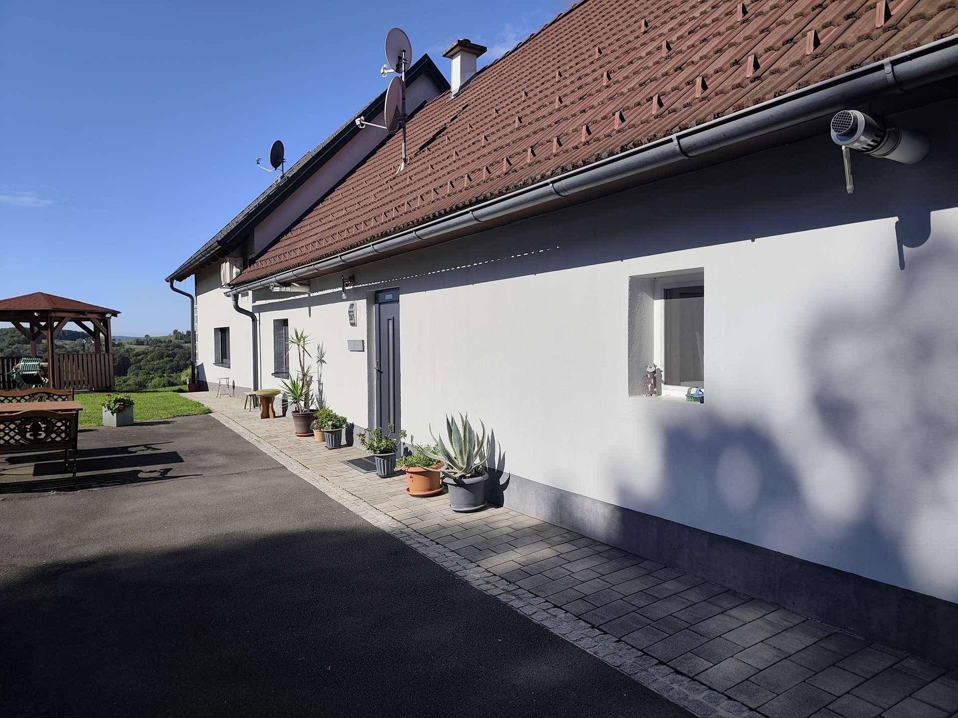 Seitenansicht eines Hauses mit gepflasterter Auffahrt, Topfpflanzen und einem Gartenpavillon im Hintergrund.