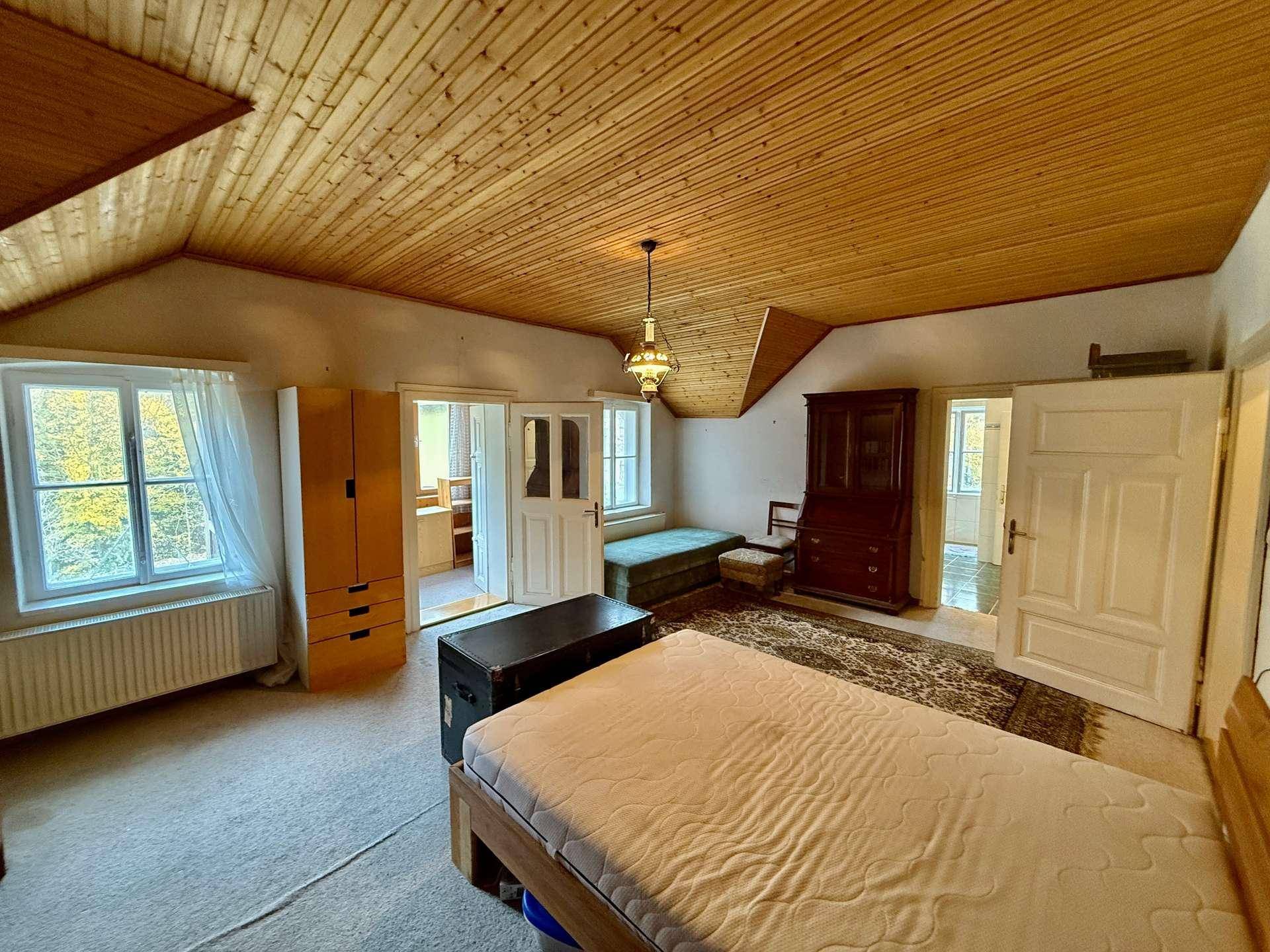 Schlafzimmer mit schräger Holzdecke, Teppichboden und Blick ins Grüne.