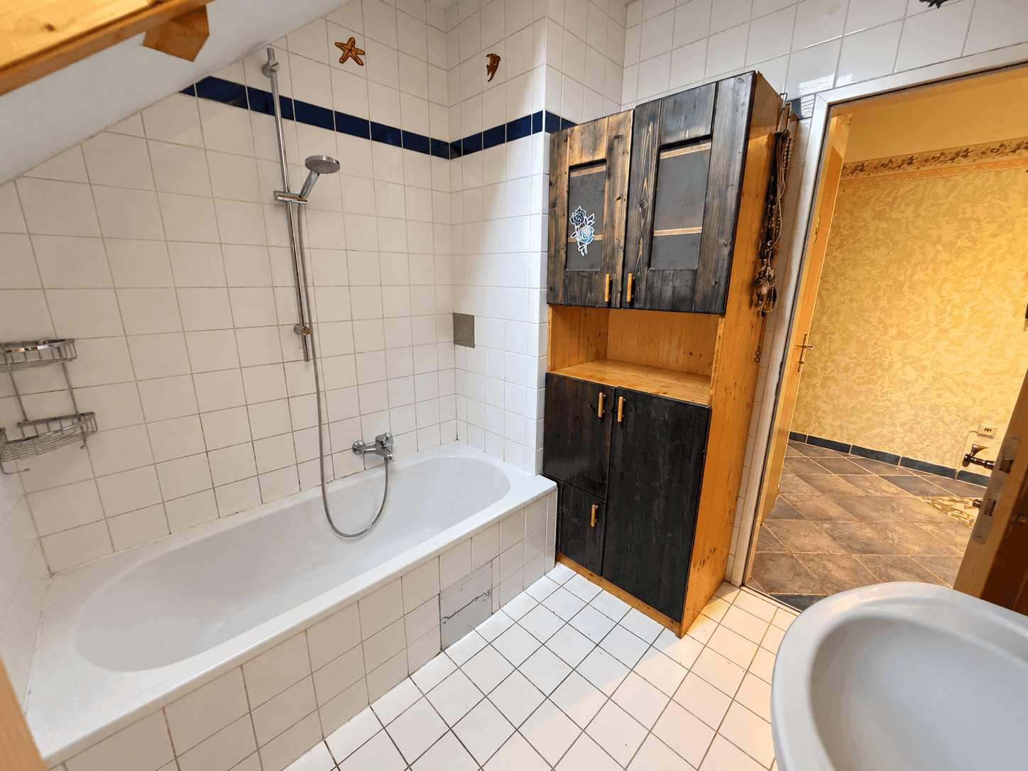 Badezimmer mit Badewanne, weißen Fliesen und Holzschrank unter Dachschräge.