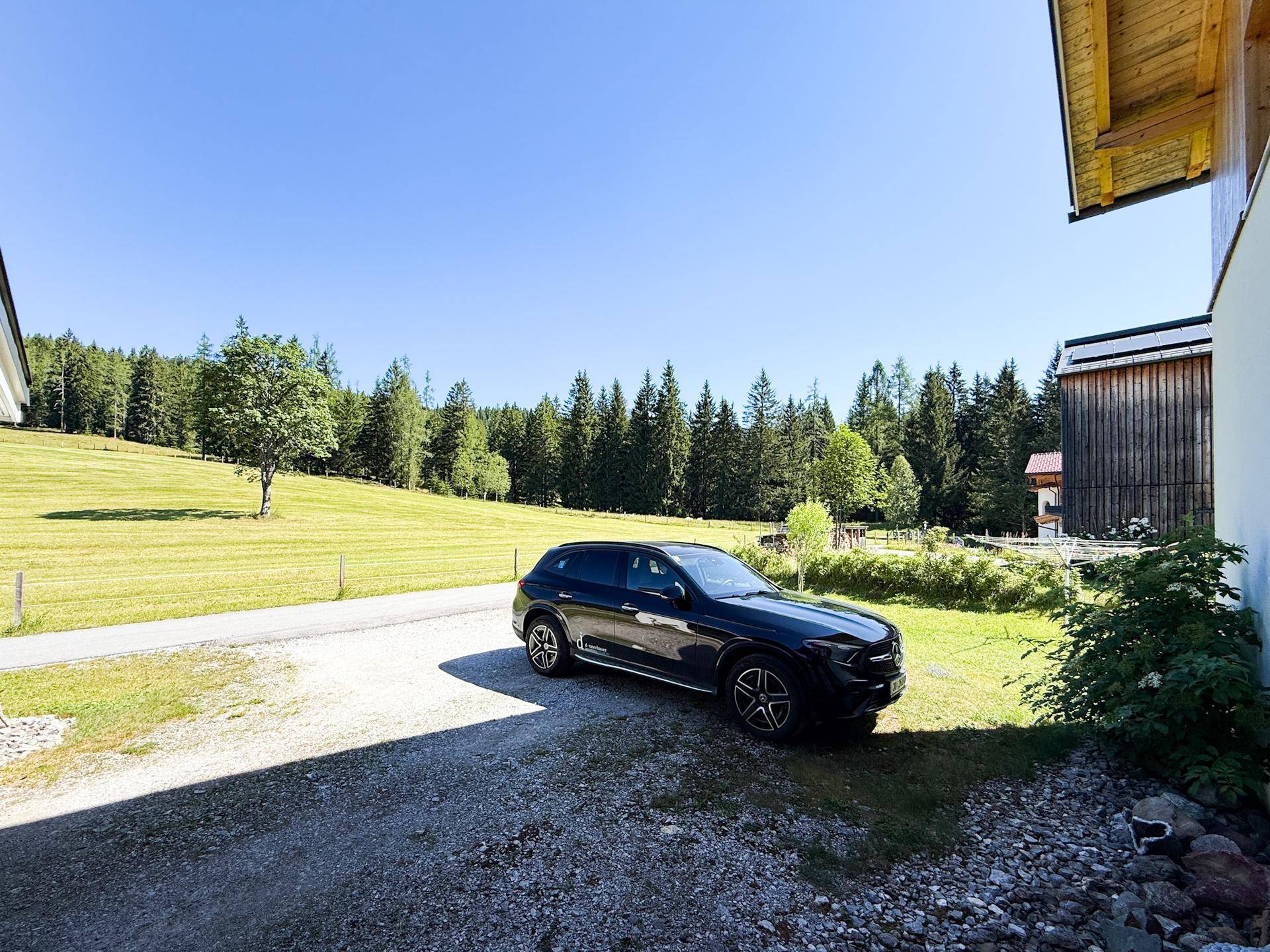 Außenparkplatz auf Schotterweg neben dem Haus, umgeben von grünen Wiesen und Wald.