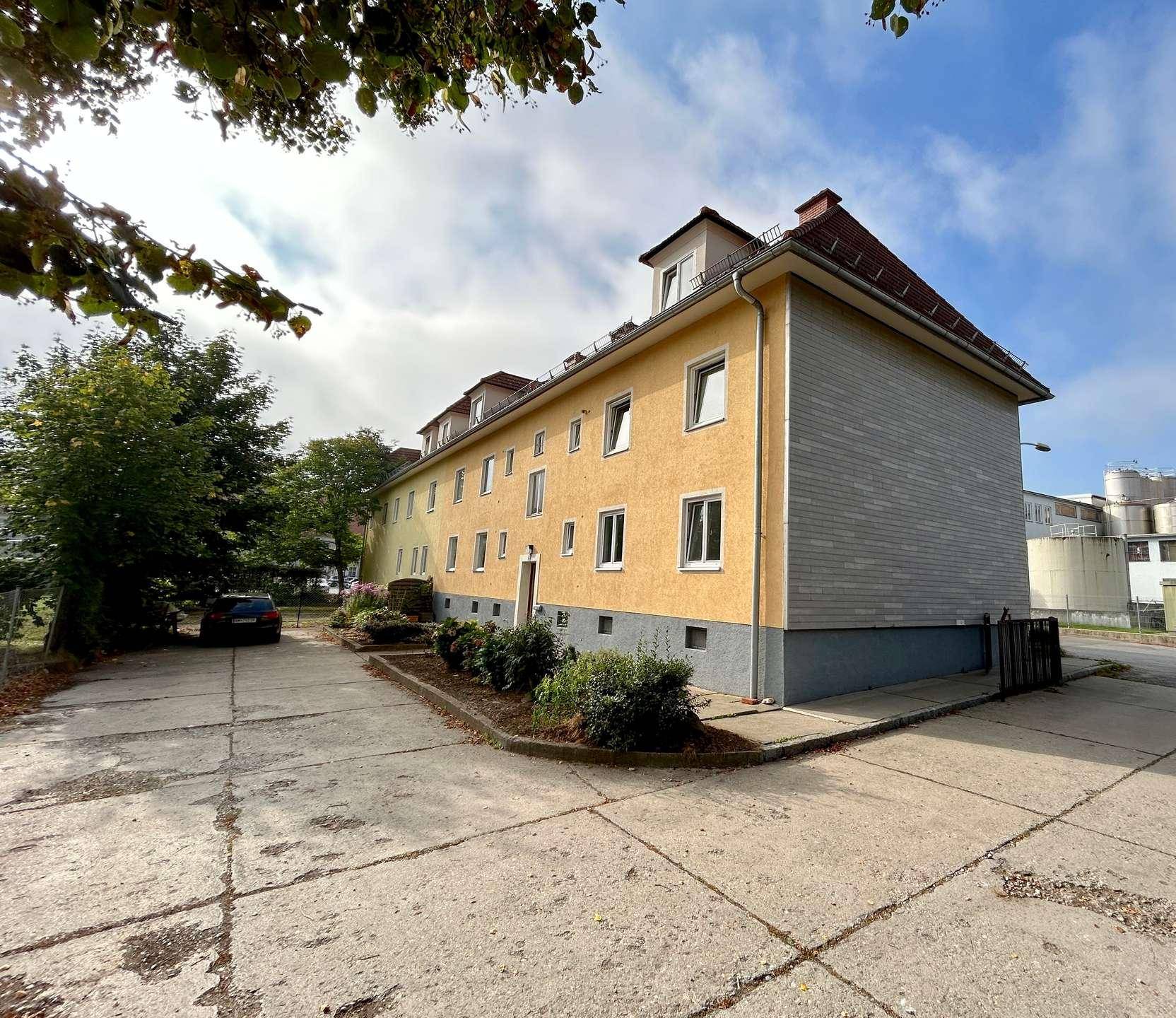 Mehrfamilienhaus mit gelber Fassade, Satteldach und Parkplätzen im Freien, umgeben von Bäumen.