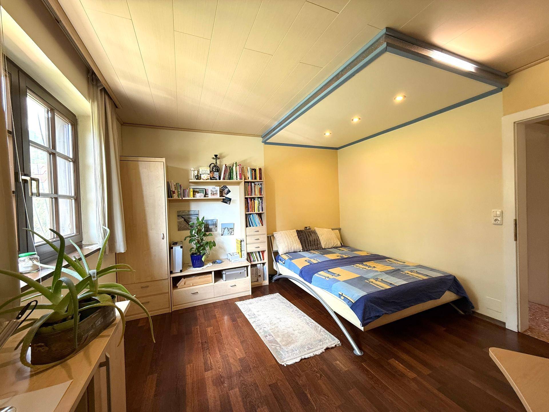 Helles Schlafzimmer mit Einzelbett, Bücherregal und Holzfußboden, ideal als Kinder- oder Gästezimmer.