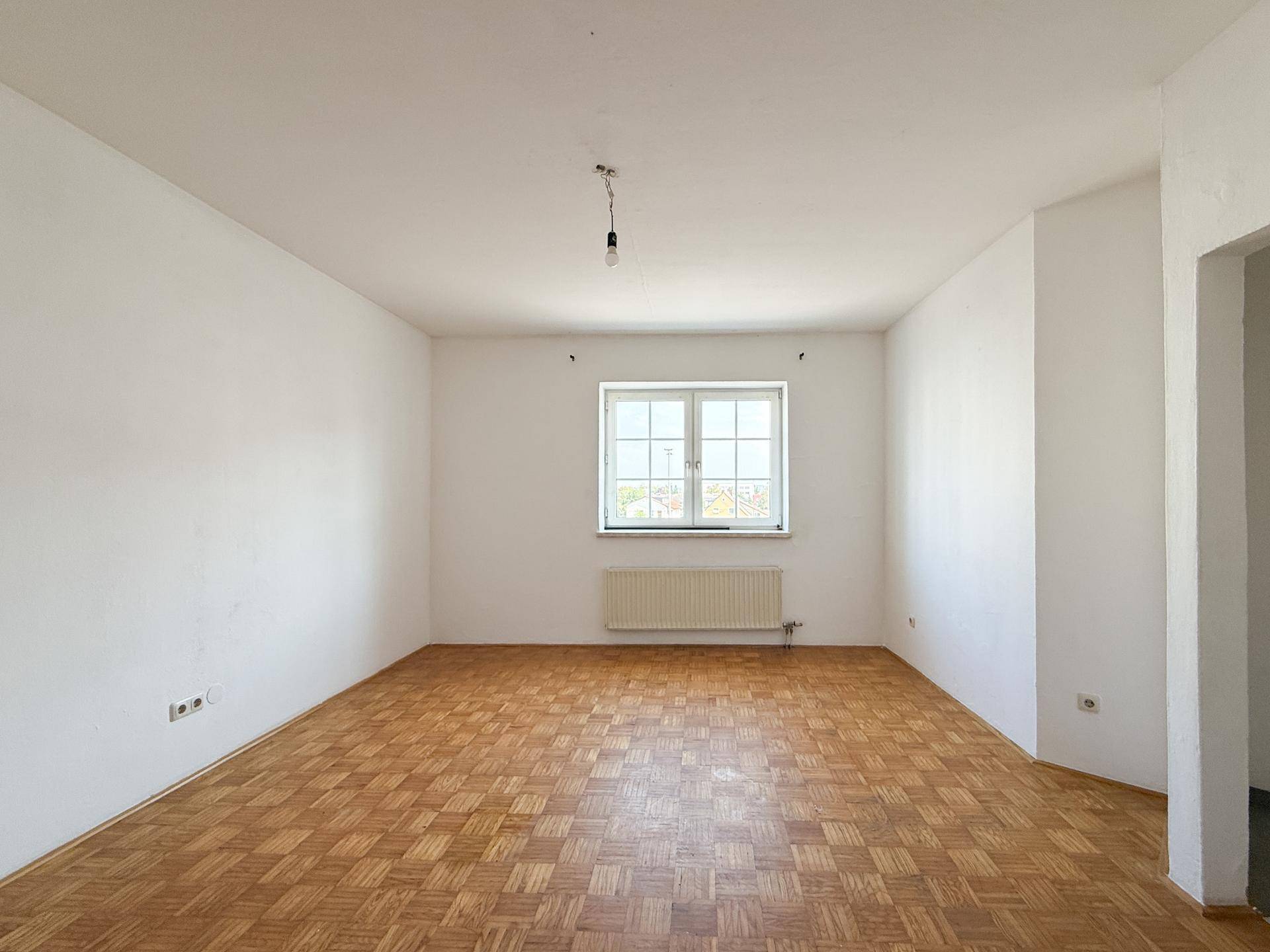 Leeres Wohnzimmer mit Parkettboden und Heizkörper unter dem Fenster, bereit zur individuellen Gestaltung.