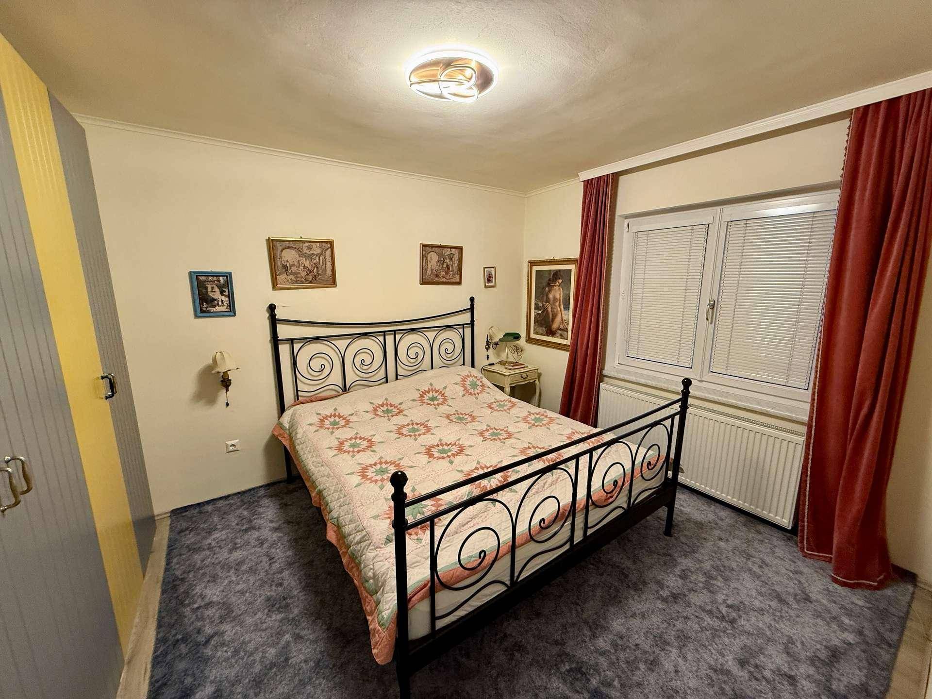 Schlafzimmer mit schmiedeeisernem Doppelbett, blauem Teppichboden und Fenster mit Jalousien.
