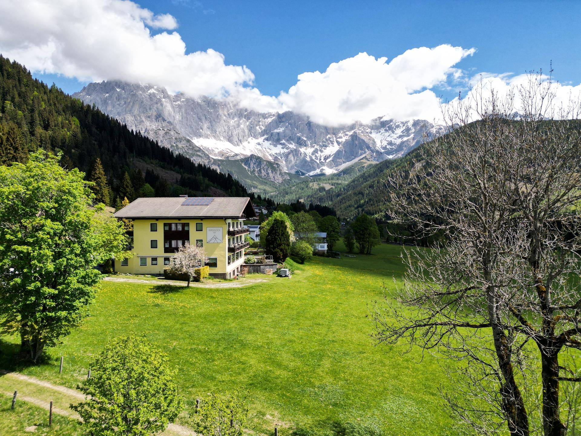 Panoramablick auf ein gelbes Gebäude in einer weiten grünen Landschaft mit majestätischen Bergen im Hintergrund.
