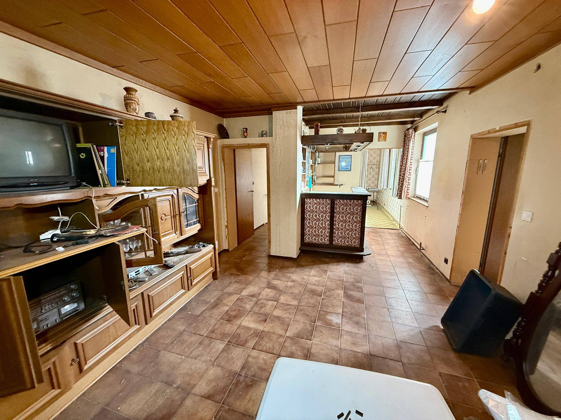 Wohnbereich mit Fliesenboden, Holzdecke und einer kleinen Bar, renovierungsbedürftig.