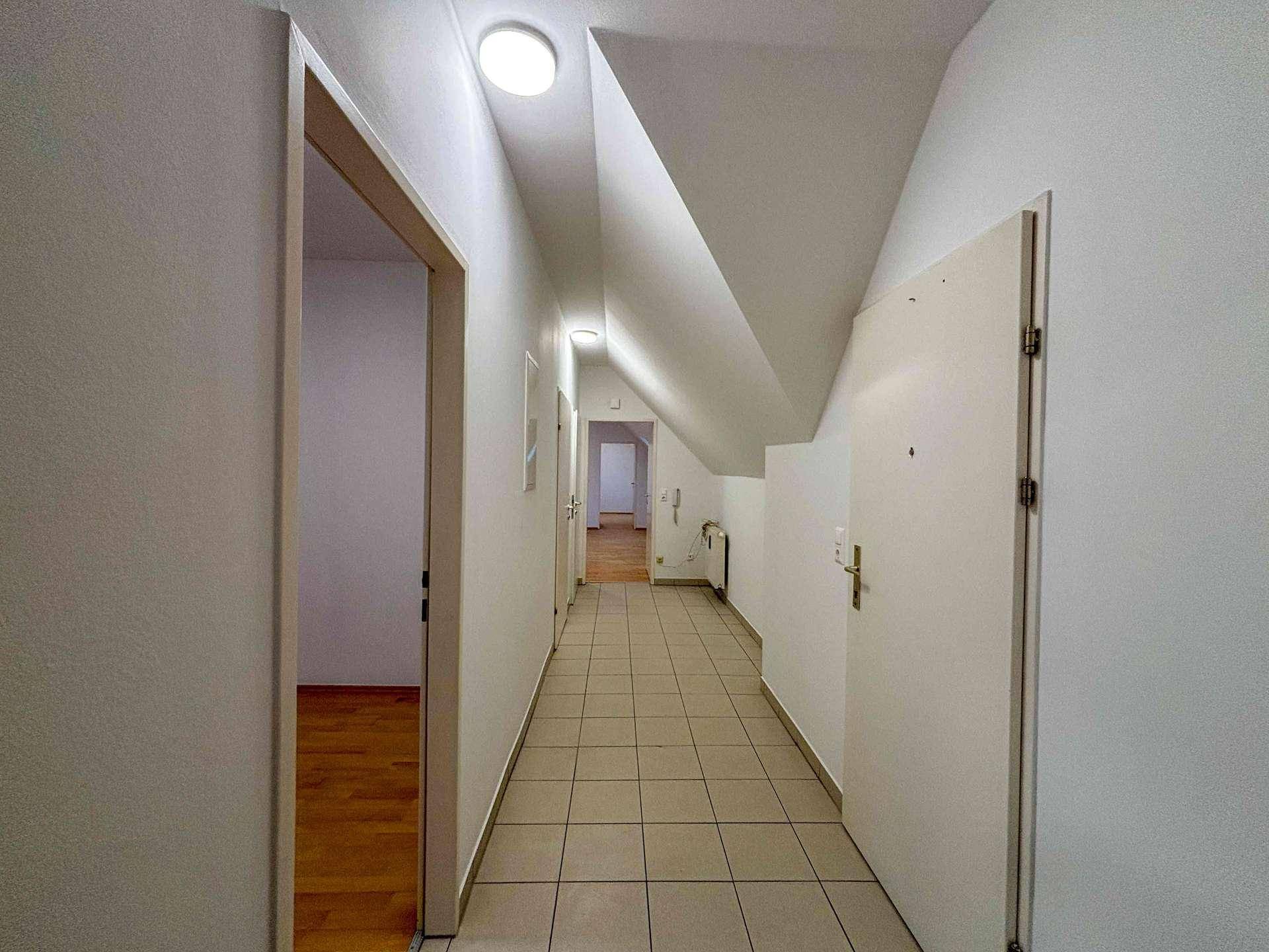 Langer Flur mit hellen Fliesen, weißen Wänden und schräger Decke im Dachgeschoss.