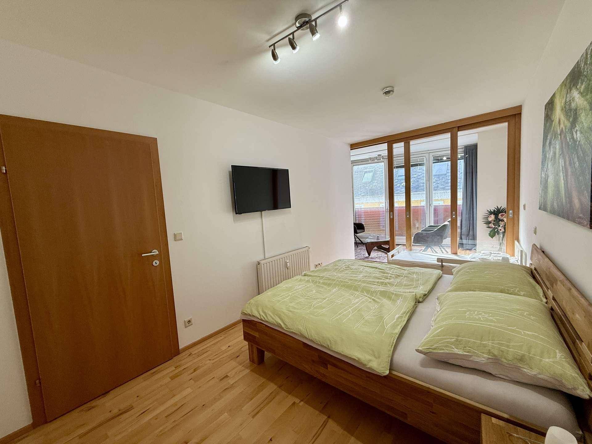 Helles Schlafzimmer mit Holzbett, Fernseher und Zugang zu einem sonnigen Balkon.