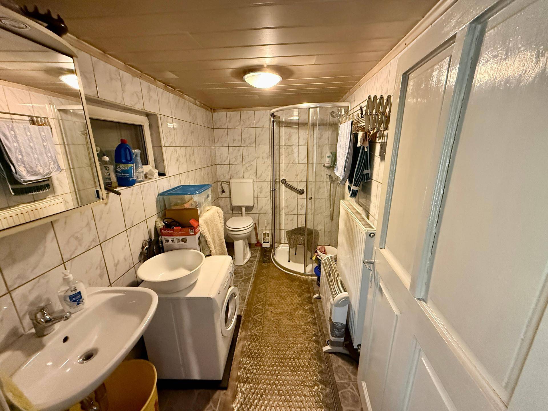 Kleines Badezimmer mit Fliesen, Dusche, WC, Waschmaschine und Haltegriffen für Barrierefreiheit.